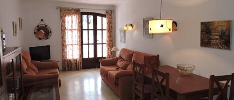 A living room with a sofa set, a TV, a dining table with chairs, and a large window with curtains.