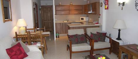 A living room with a sofa, chairs, a coffee table with fruit, and a kitchen in the background.