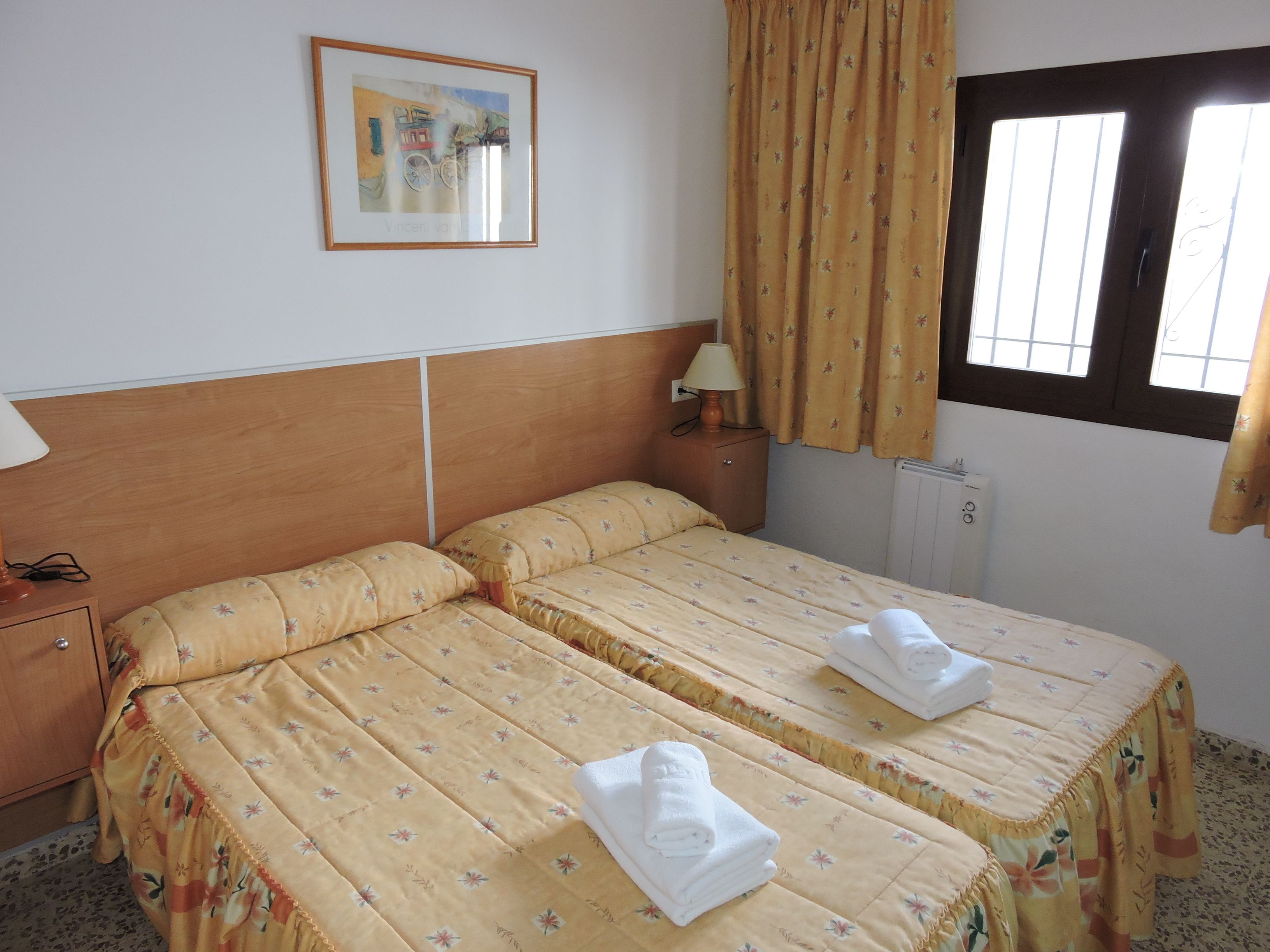 A hotel room with two beds, a wooden headboard, a nightstand, a lamp, a window with curtains, and a framed picture on the wall.