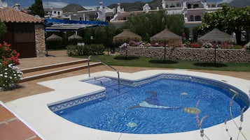 A swimming pool with a mosaic border, surrounded by a stone wall and thatched umbrellas.