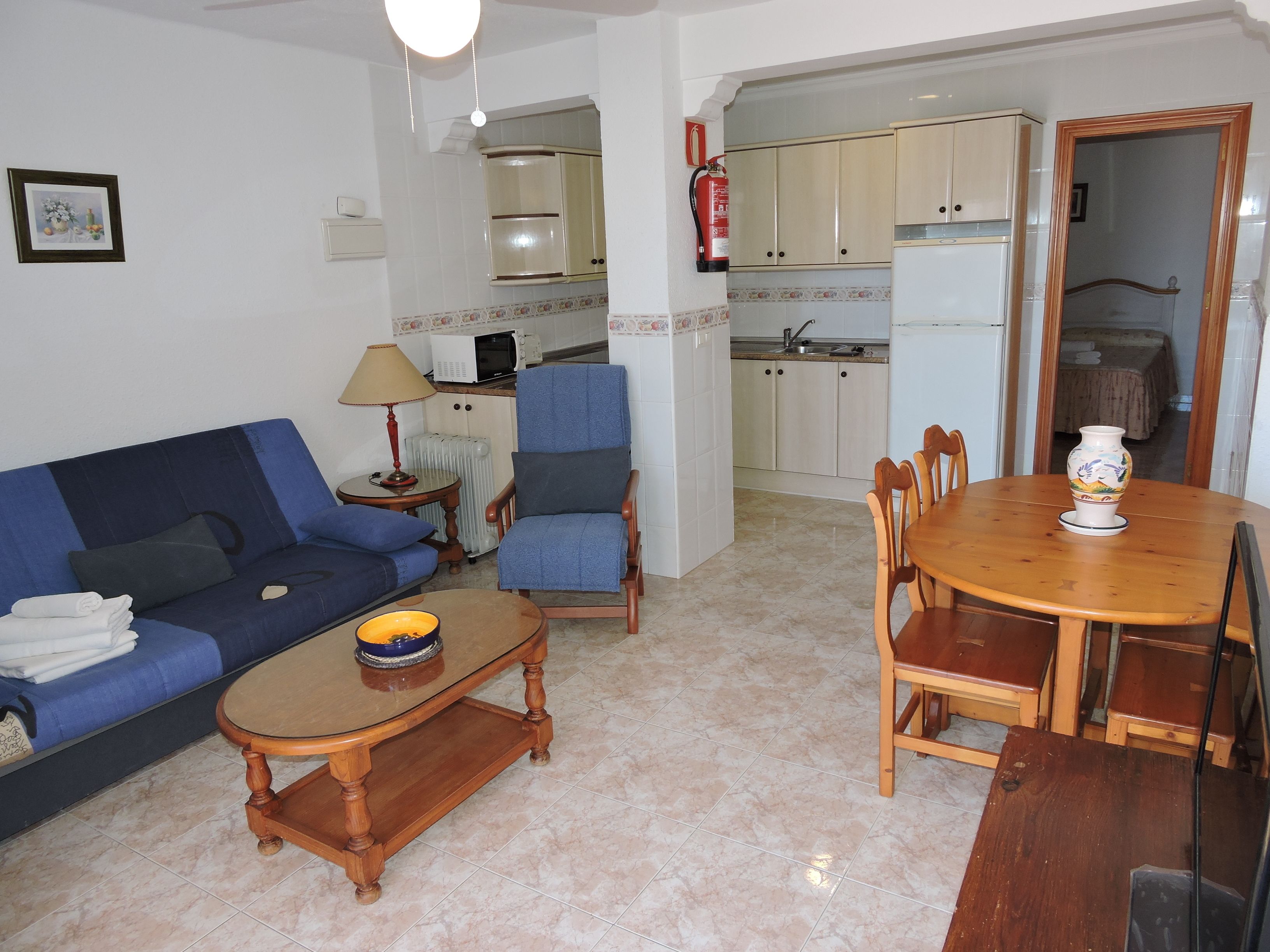 A living room with a sofa, armchair, coffee table, and dining table. There is a kitchen in the background with cabinets and a refrigerator.