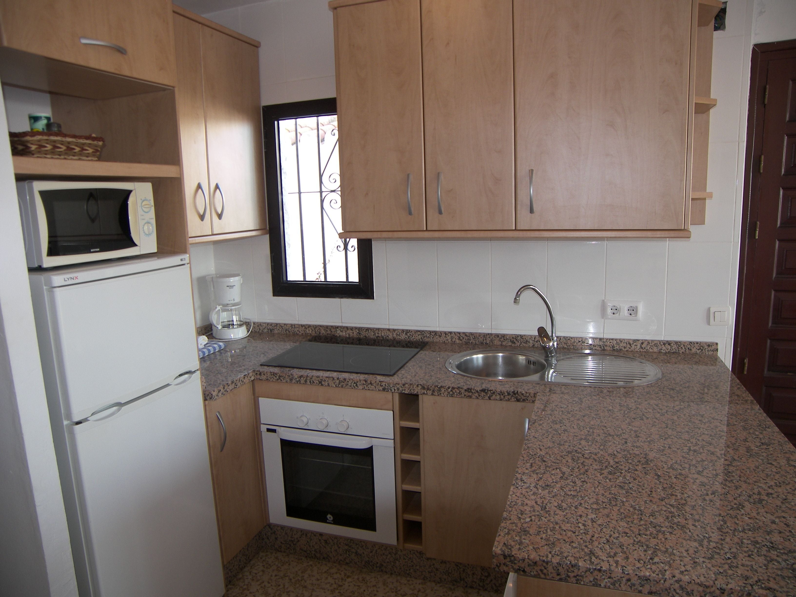 A compact kitchen with wooden cabinets, a microwave, a coffee maker, a sink, and a window with bars.
