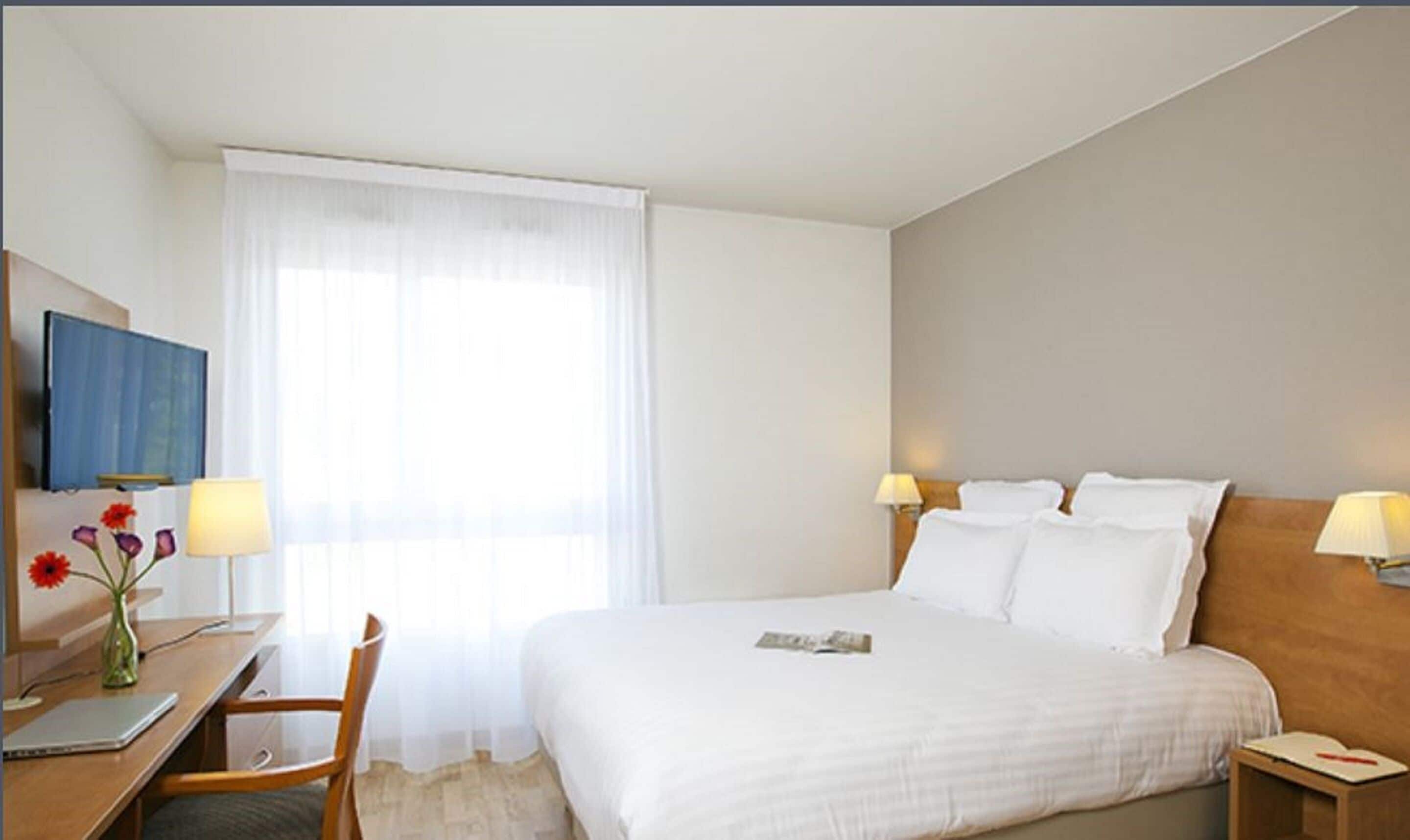 A hotel room with a bed, bedside lamps, a desk with a laptop, a chair, and a window with sheer curtains.