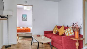 A modern room with a red sofa, a wooden coffee table, and a small side table with a vase. A bed with an orange blanket is visible in the background.
