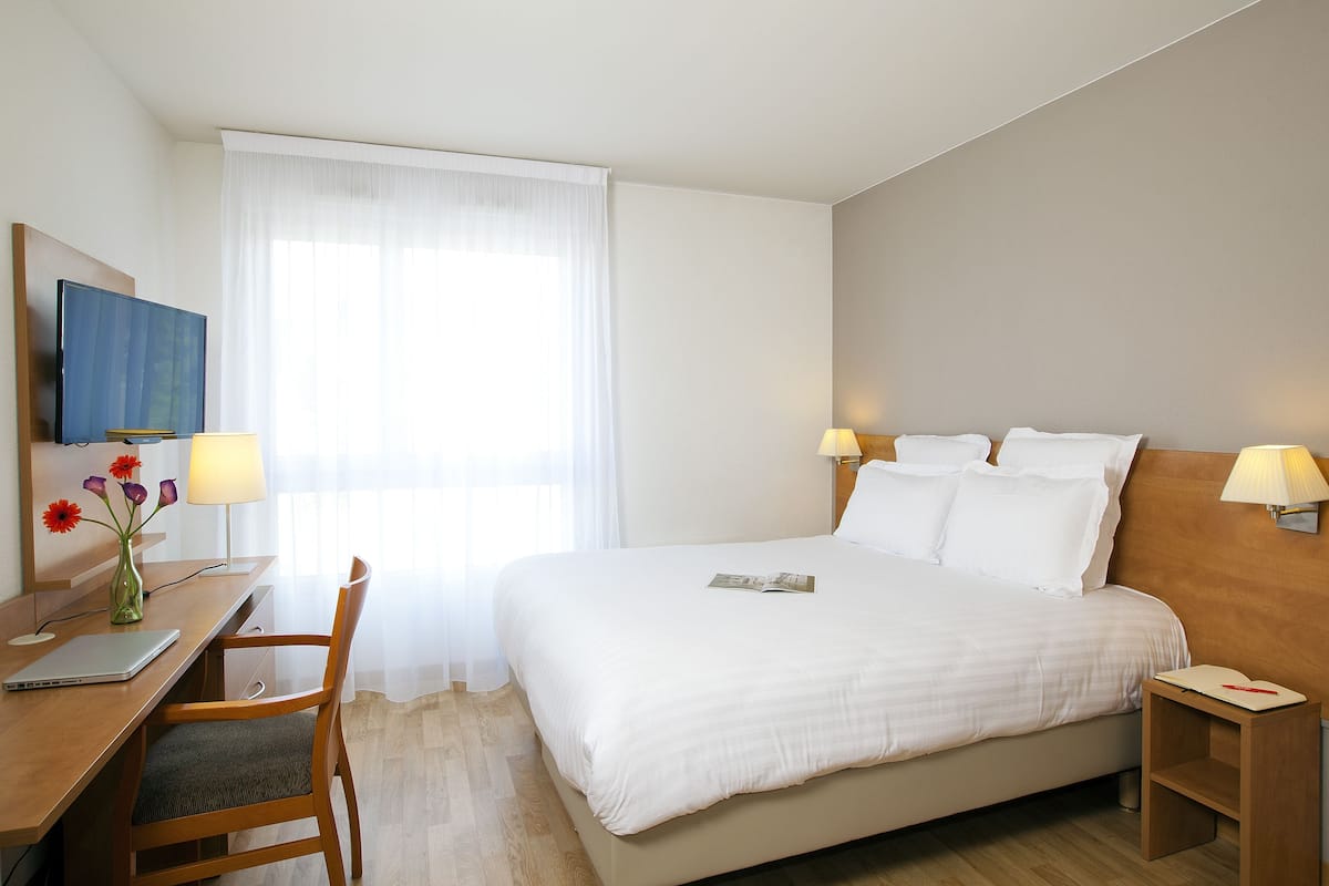 Ein Hotelzimmer mit einem Bett, einem Schreibtisch mit Laptop, einem Sessel und zwei Nachttischlampen.