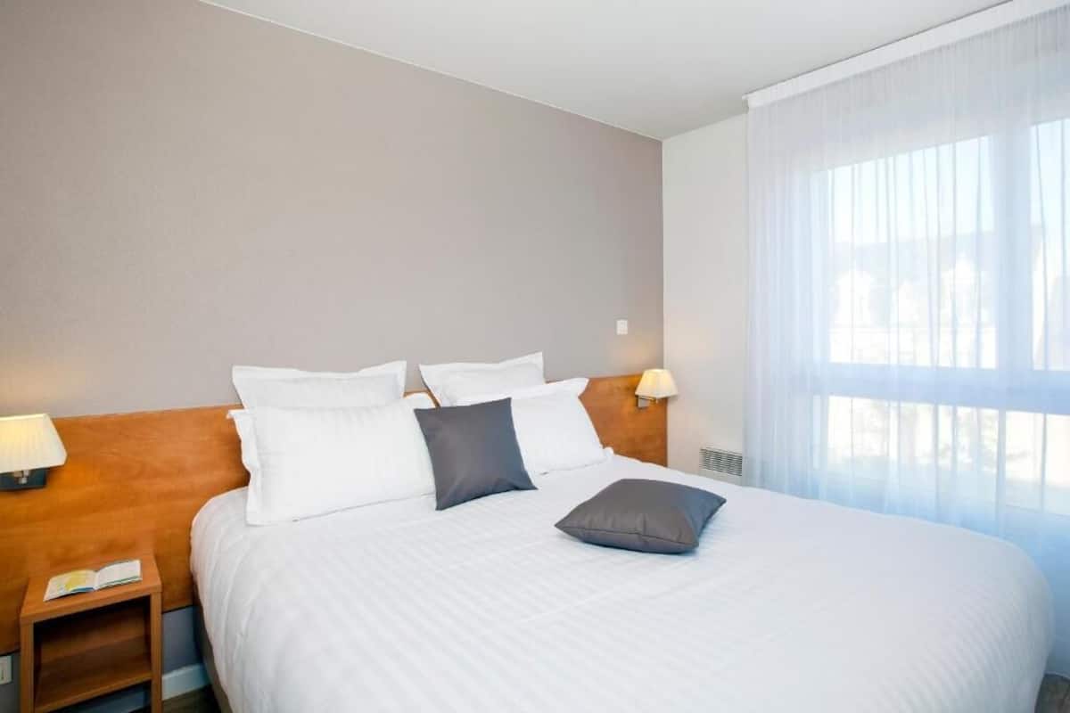 A hotel room with a large bed, two bedside tables, a nightstand, a window with sheer curtains, and a headboard.