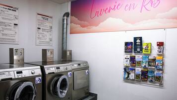 A laundry area with industrial washers and dryers, informational posters on the wall, and a bulletin board with various items pinned to it.