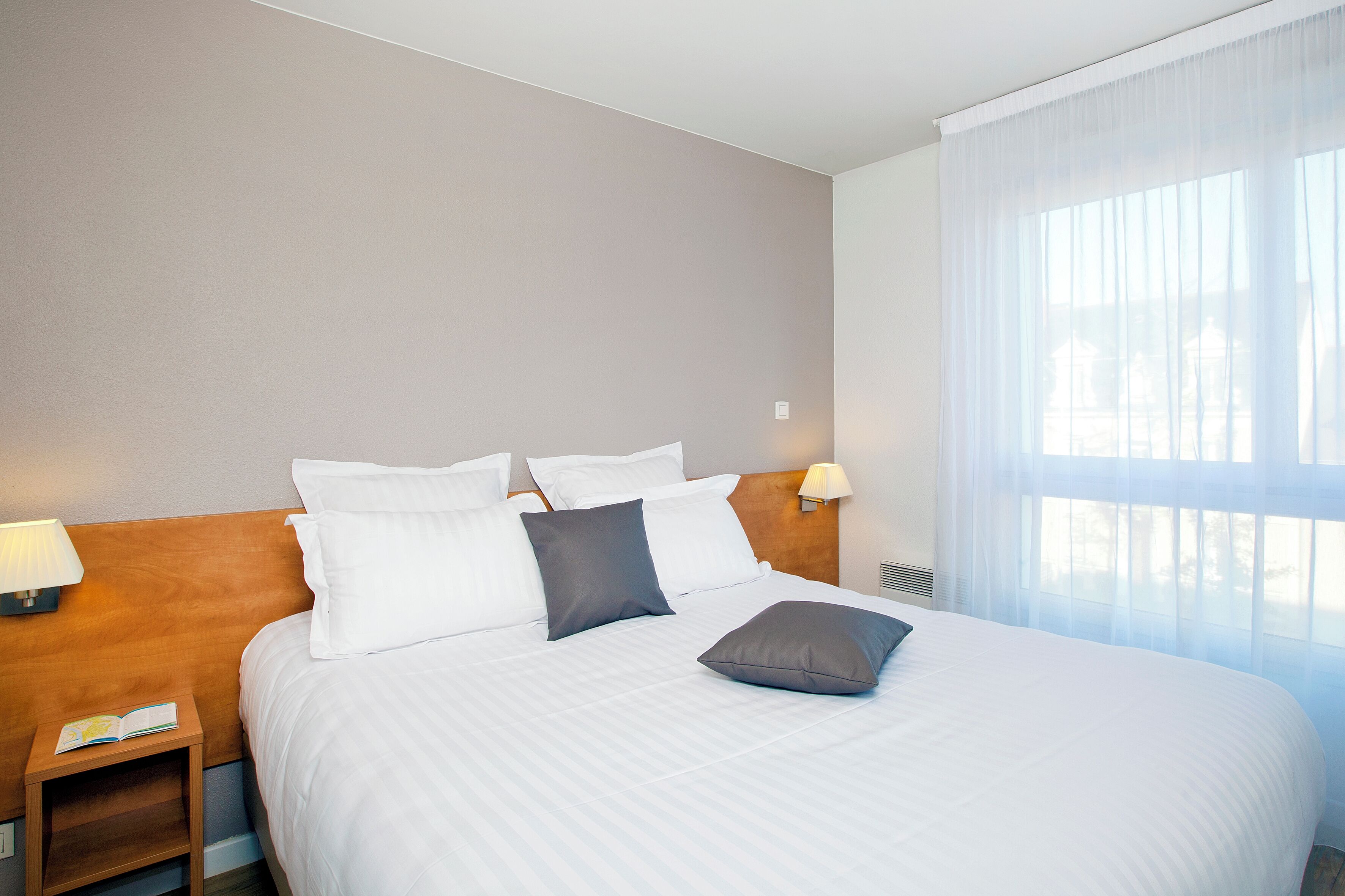 A hotel room with a neatly made bed, a bedside table with a lamp, and a window with sheer curtains.