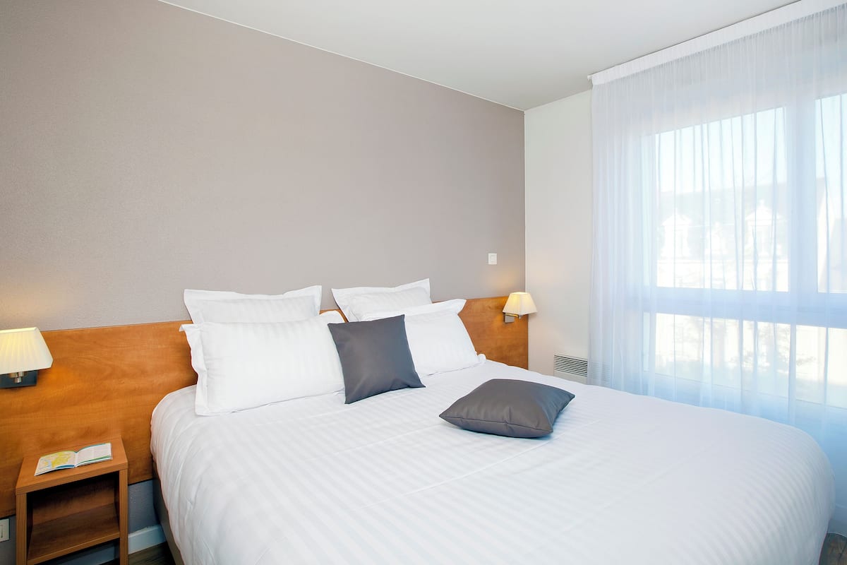 A hotel room with a neatly made bed, a bedside table with a lamp, and a window with sheer curtains.