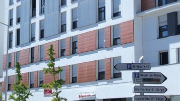 Un bâtiment moderne à plusieurs étages, doté d’une façade blanche et de touches de couleur rouge. Des panneaux de signalisation indiquent les lieux d’intérêt à proximité, tels que les « Berges de Maine » et l’« Université S’ Serge ».