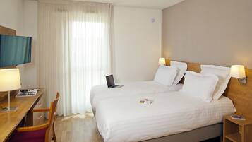 A hotel room with a bed, a desk with a laptop, a chair, a TV, and a window with curtains.