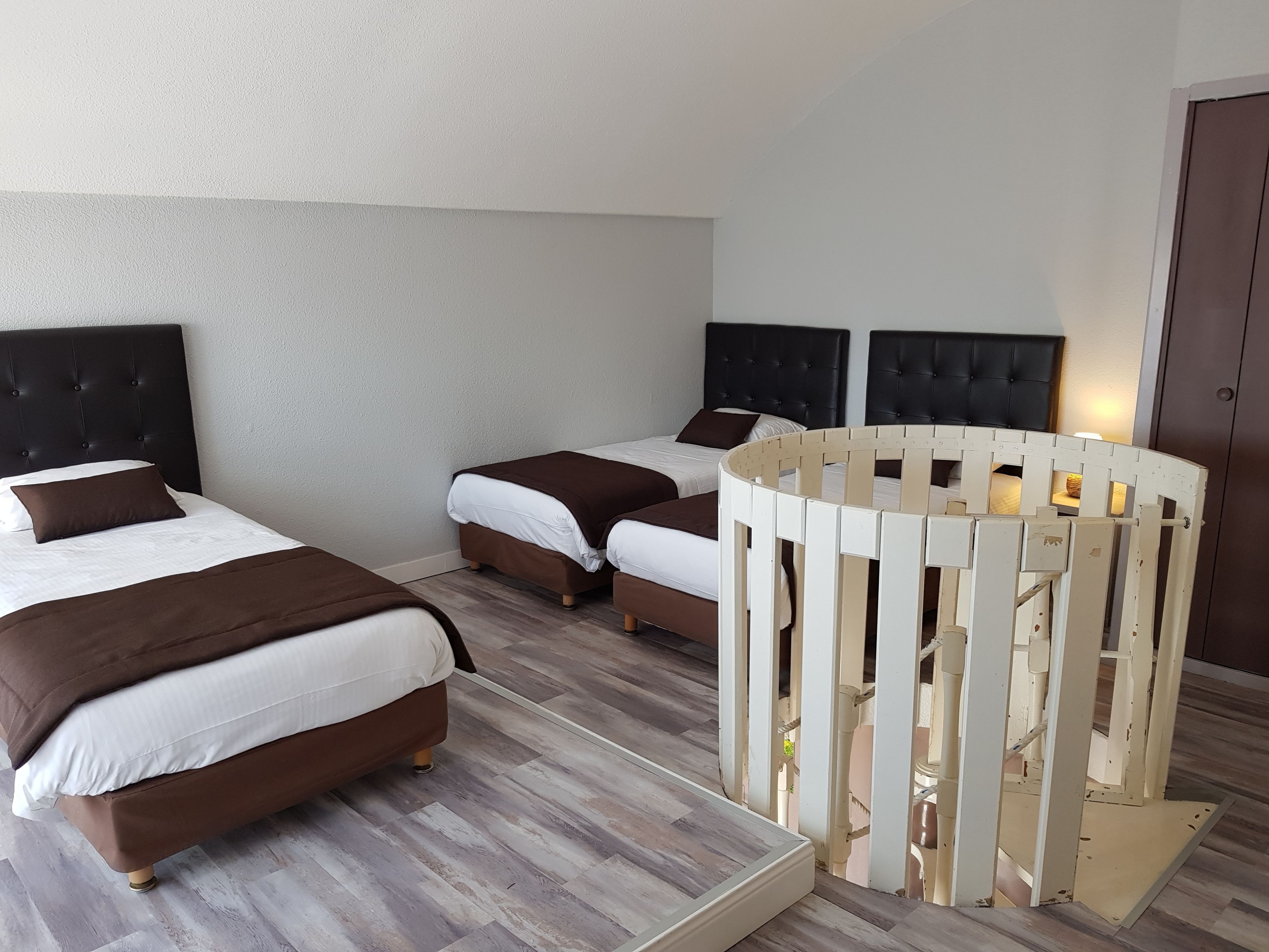 Une chambre d’hôtel avec deux lits, chacun doté d’une tête de lit sombre et de draps blancs. Un berceau blanc est placé dans un coin de la pièce.