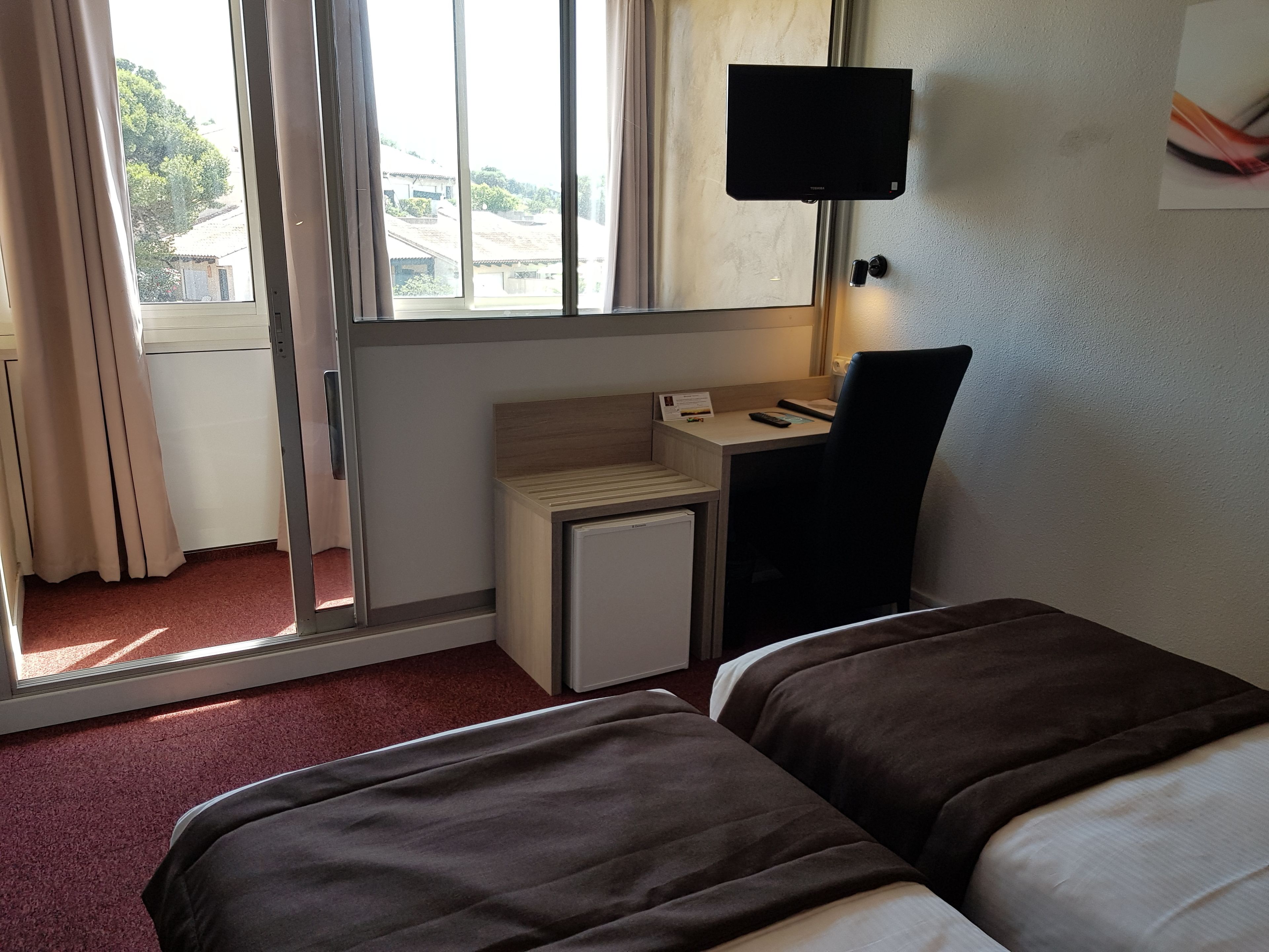 A hotel room with two beds, a desk, a small refrigerator, a television, and a window with curtains.