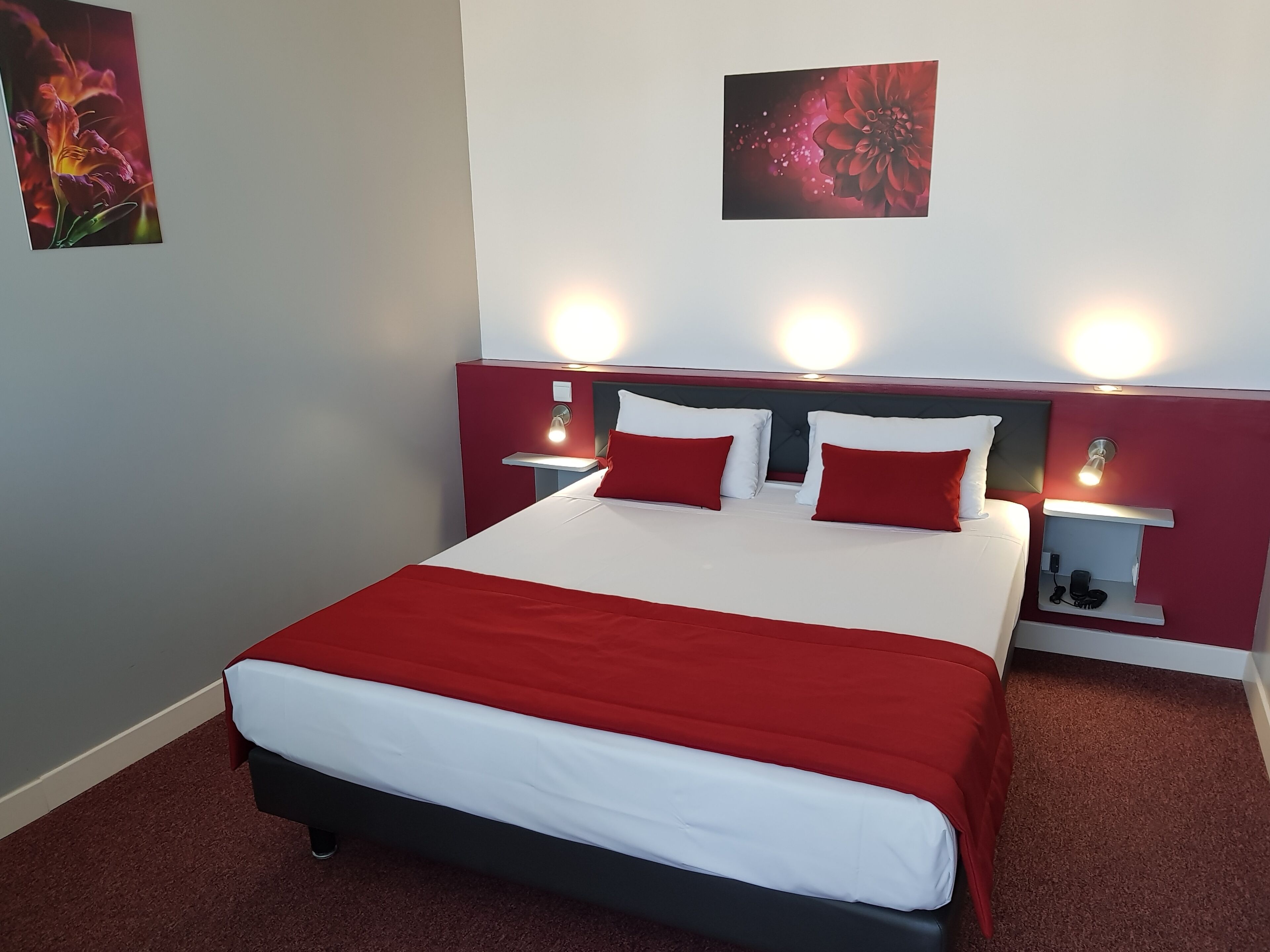Quarto de hotel com cama, roupa de cama vermelha e branca, duas mesas de cabeceira com luminárias e duas luminárias fixadas na parede.