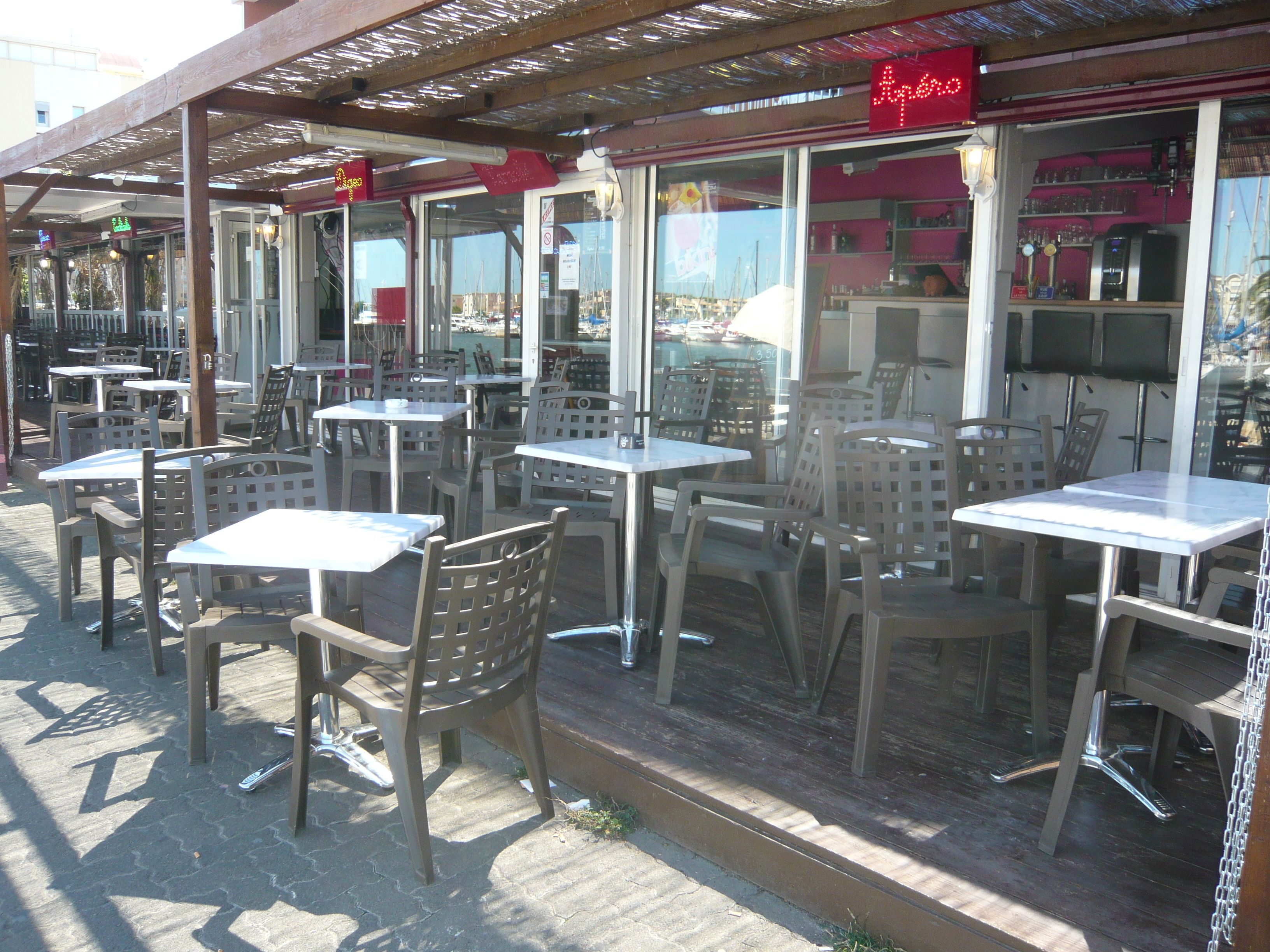 En udendørs café med borde og stole, en træpergola og et rødt neonskilt.