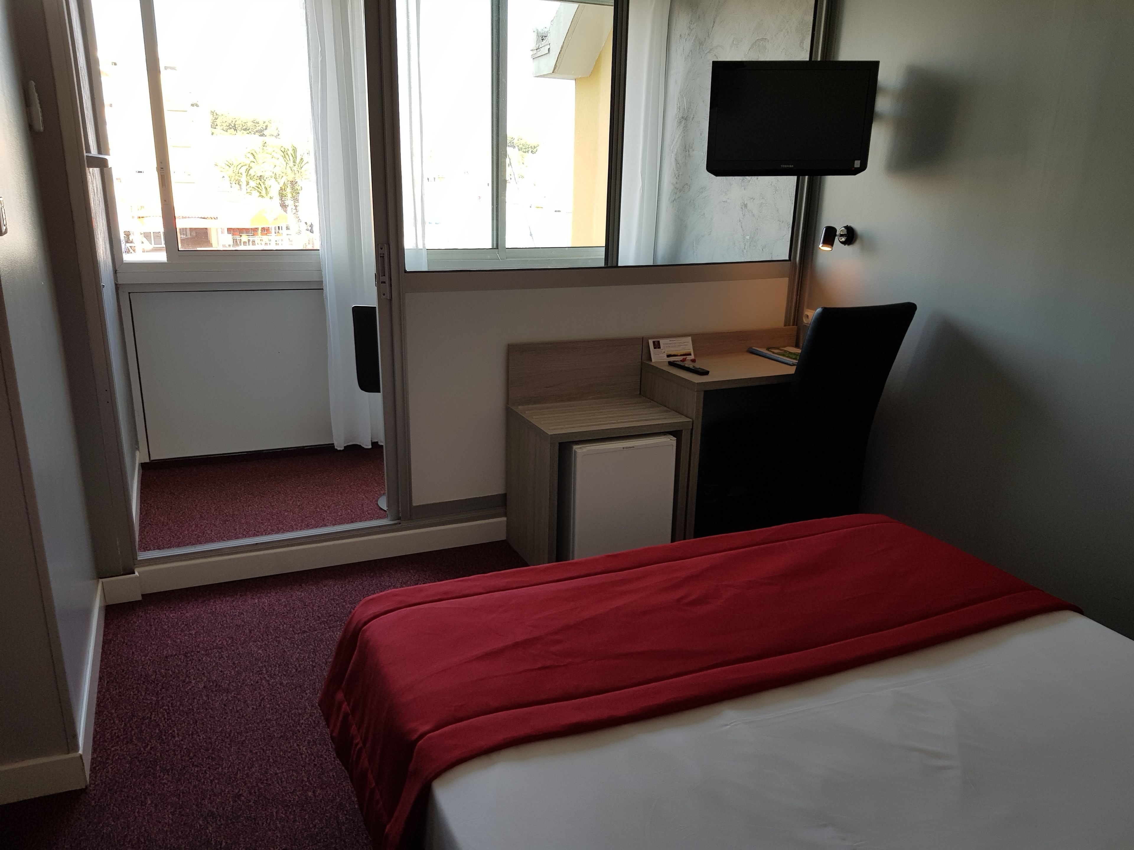 Una habitación de hotel con cama, un escritorio pequeño, televisión y ventana con cortinas.