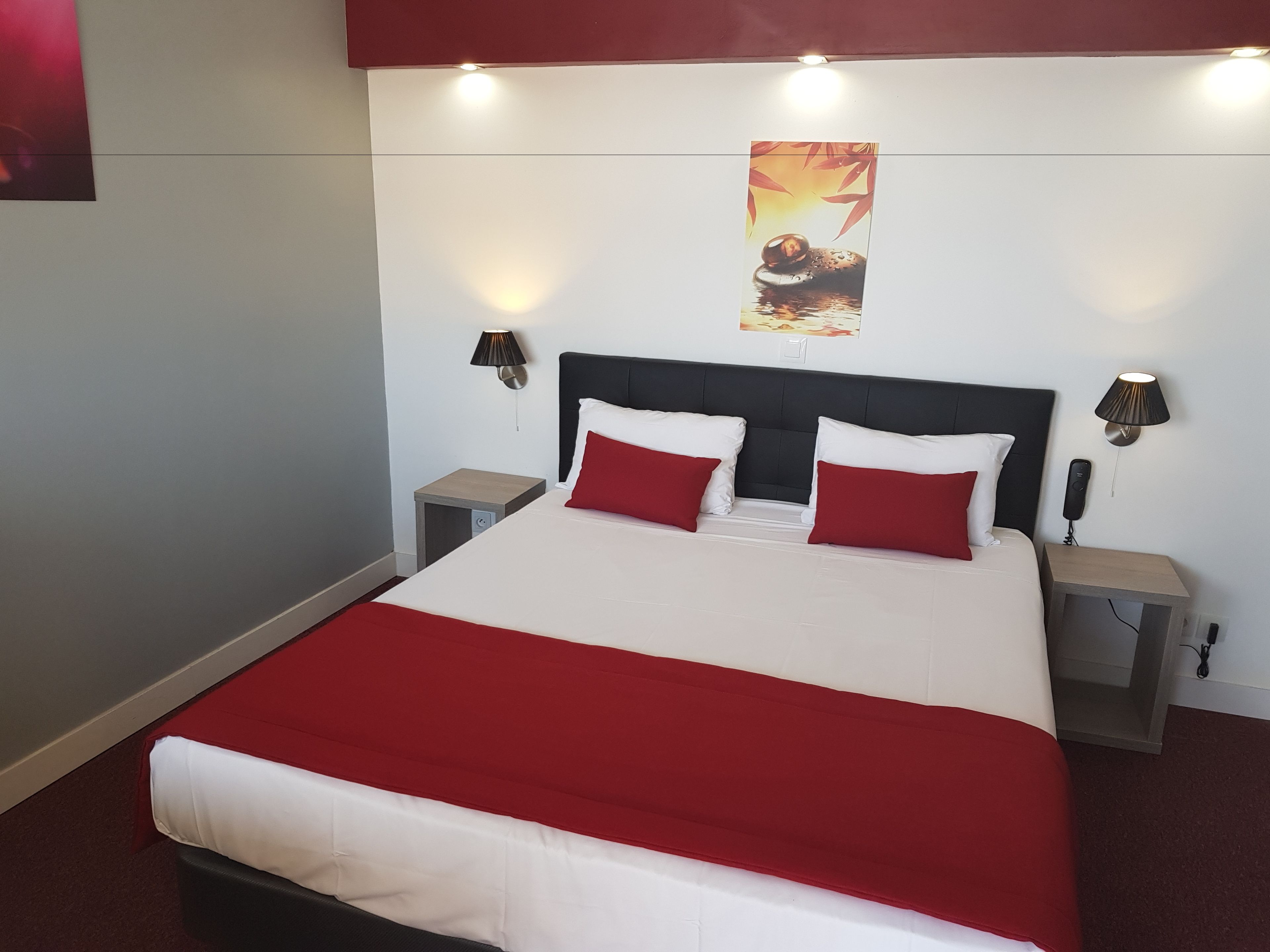 A neatly made bed with a red and white bedspread, two red pillows, and a black headboard. There are two bedside tables with lamps, and a wall-mounted light fixture above the bed.