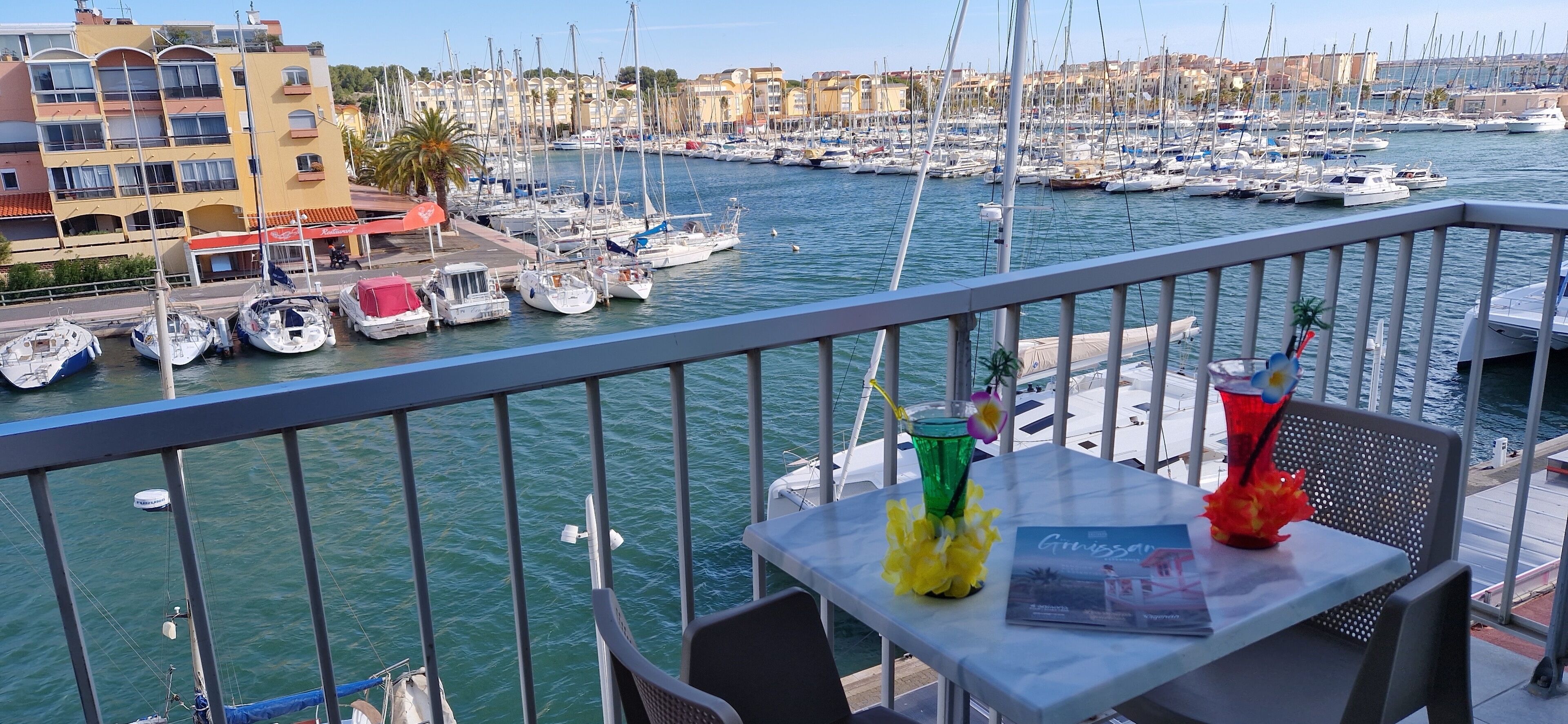 Un balcón con mesa, sillas y vistas a un puerto deportivo con barcos y edificios.