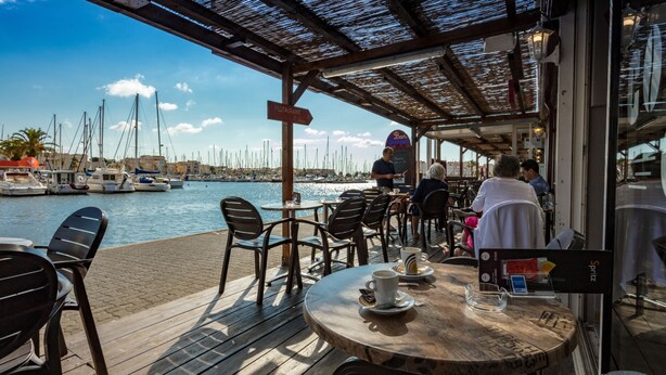 An outdoor café with wooden tables and chairs, overlooking a marina with boats.