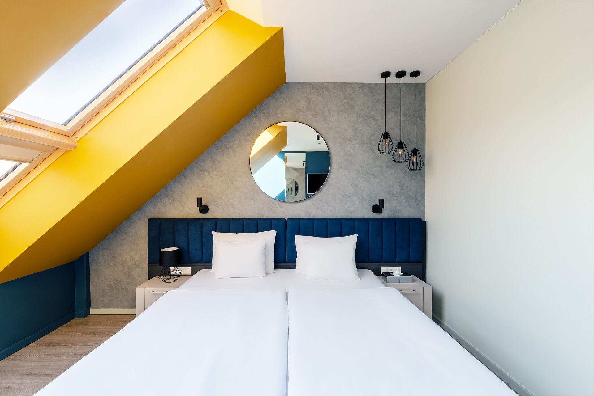 Un dormitorio con tragaluz, un espejo redondo, una cama con ropa de cama blanca y un cabecero azul.