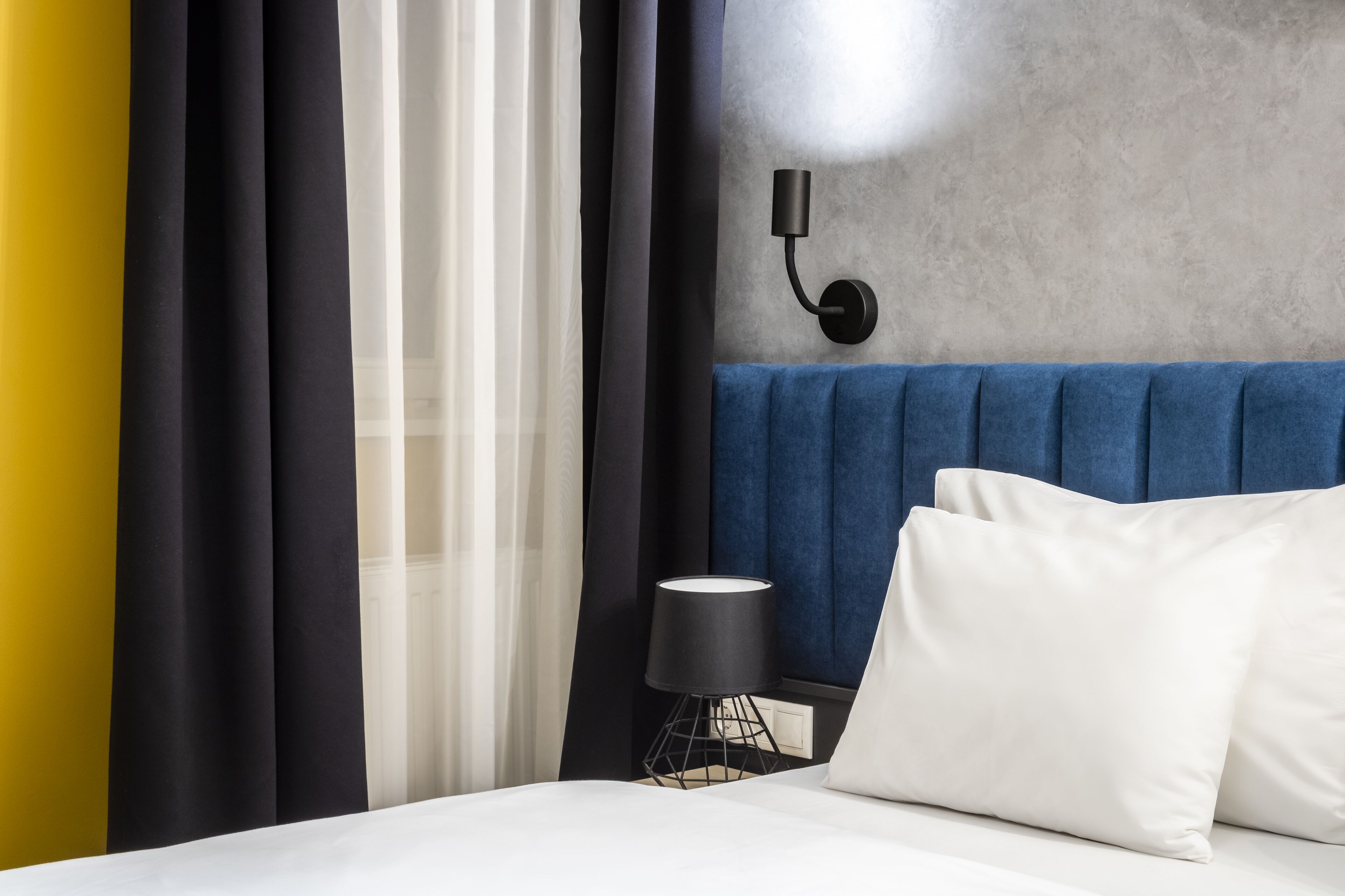 Una camera d'albergo con un letto, cuscini bianchi, una lampada nera e un faretto a parete.