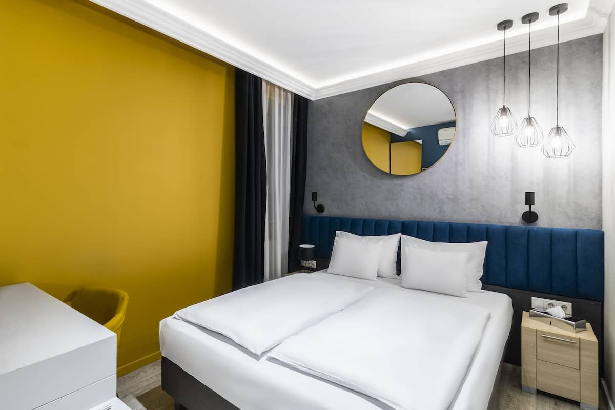 A modern hotel room with a large bed, a bedside table, a chair, and a round mirror on the wall.