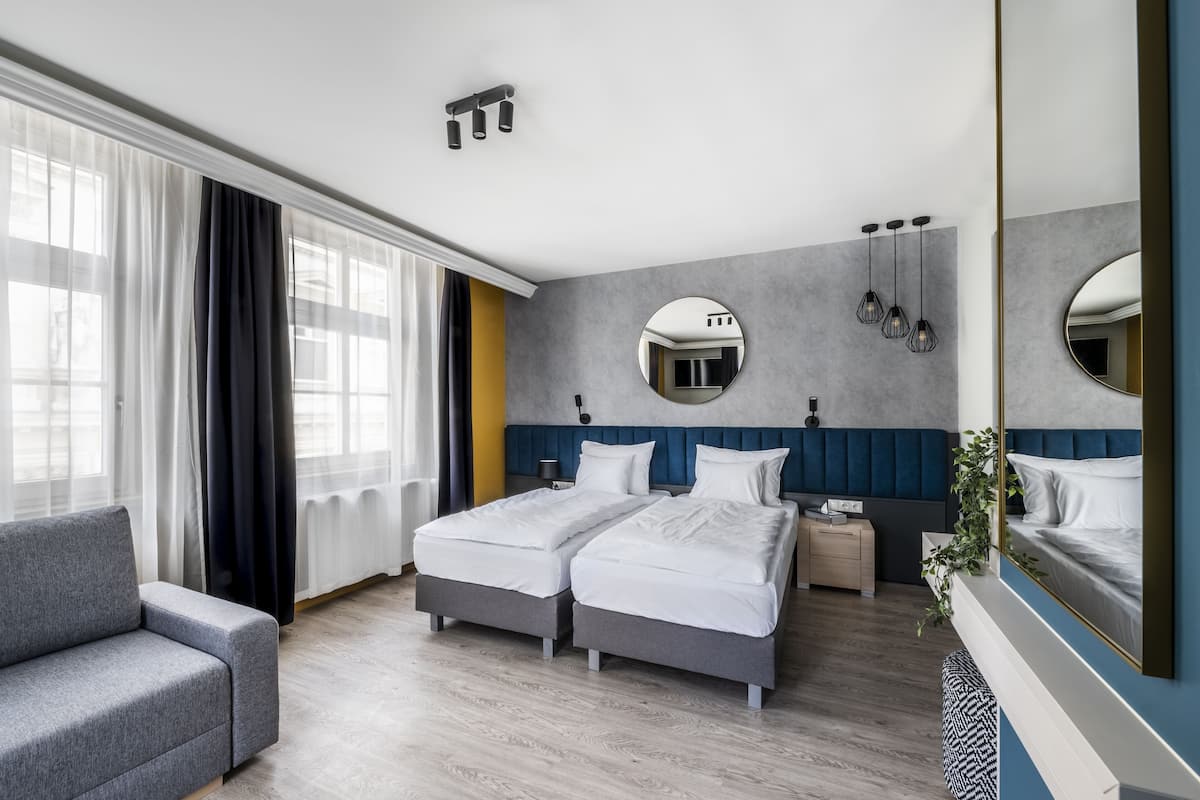 A modern hotel room with two single beds, a sofa, a bedside table, a mirror, and a large window with curtains.