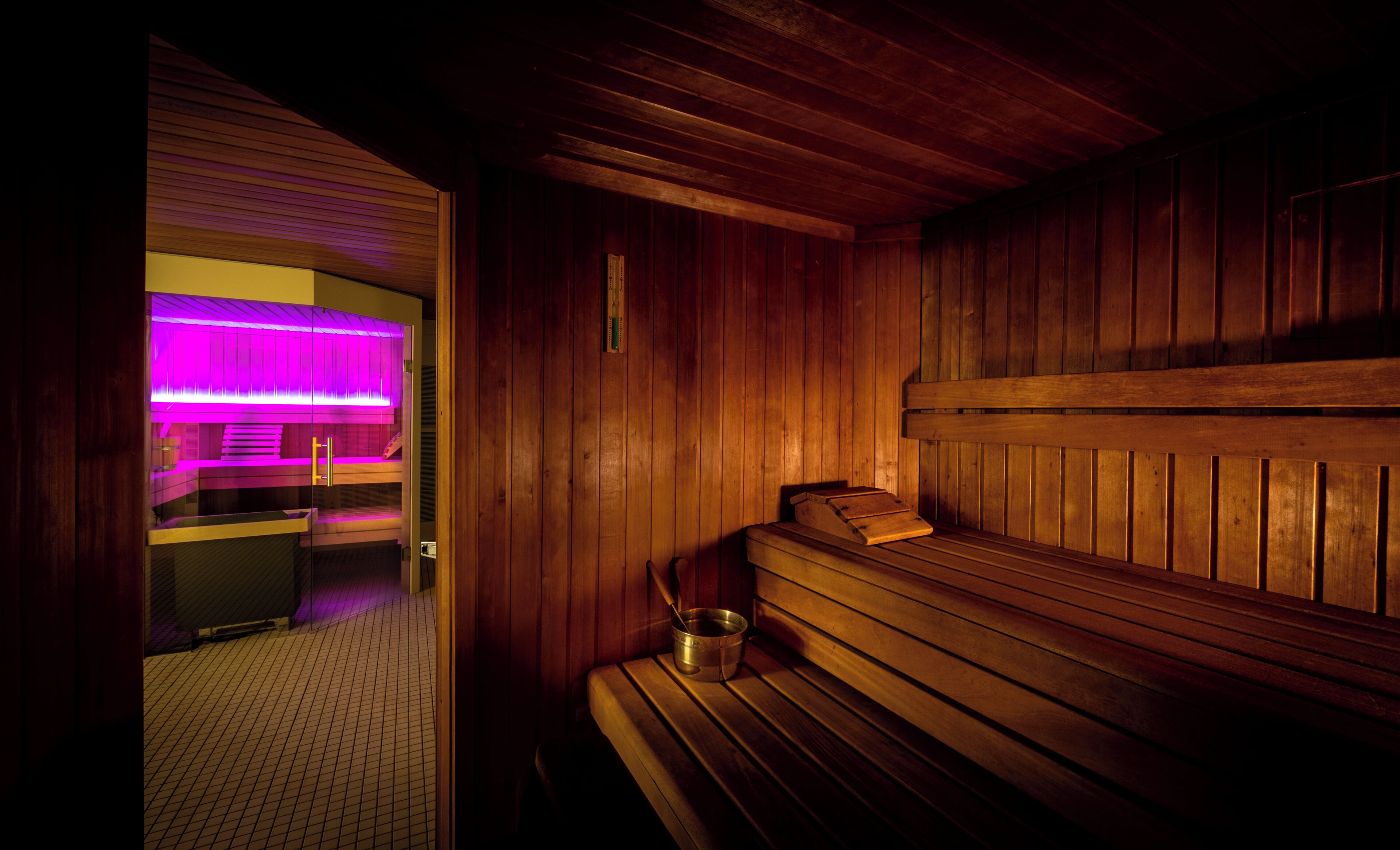 A sauna with wooden benches and a glass door leading to a changing room with a purple-lit shower area.