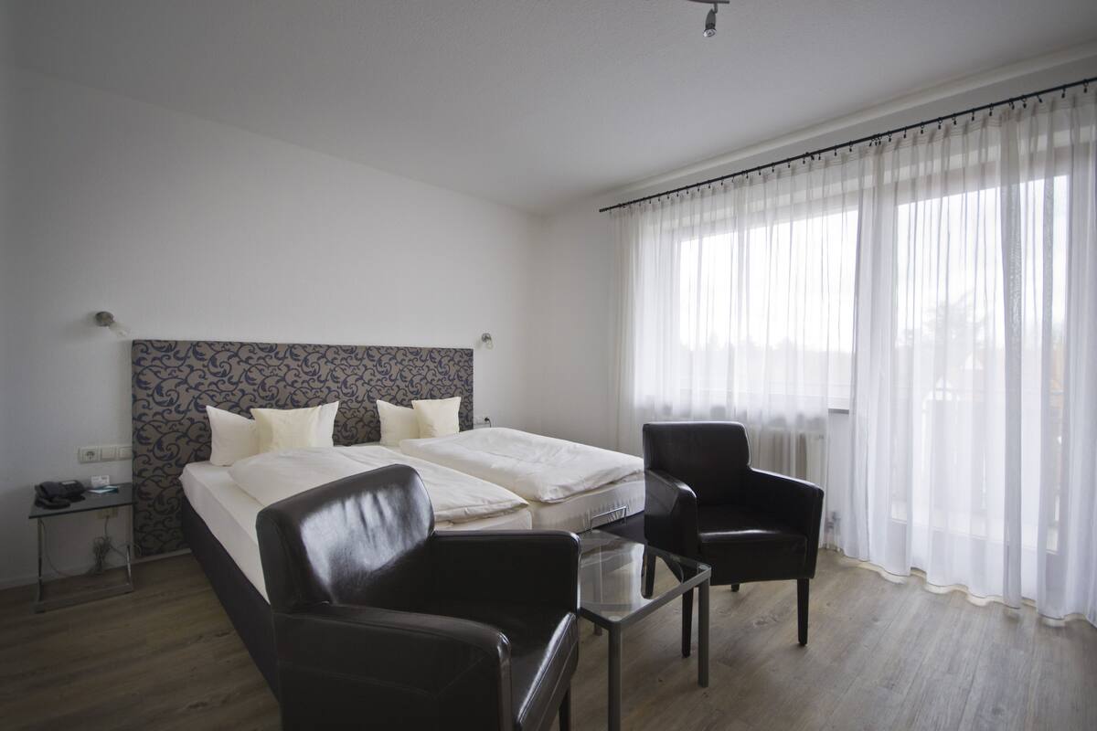 A hotel room with two beds, a small table, a chair, and a window with sheer curtains.