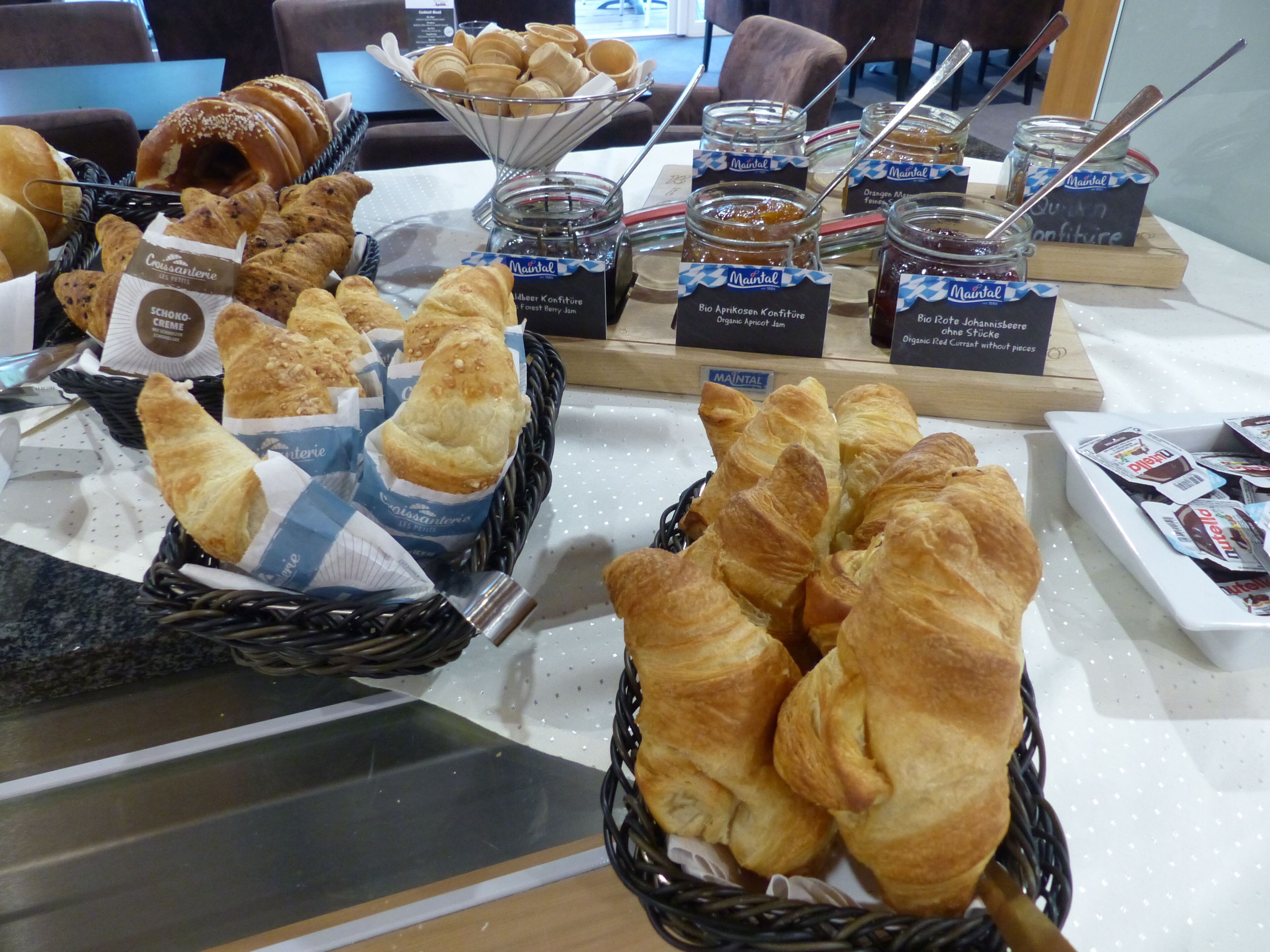 Un bufé con diversos productos horneados, incluyendo croissants y pasteles, además de frascos con conservas.