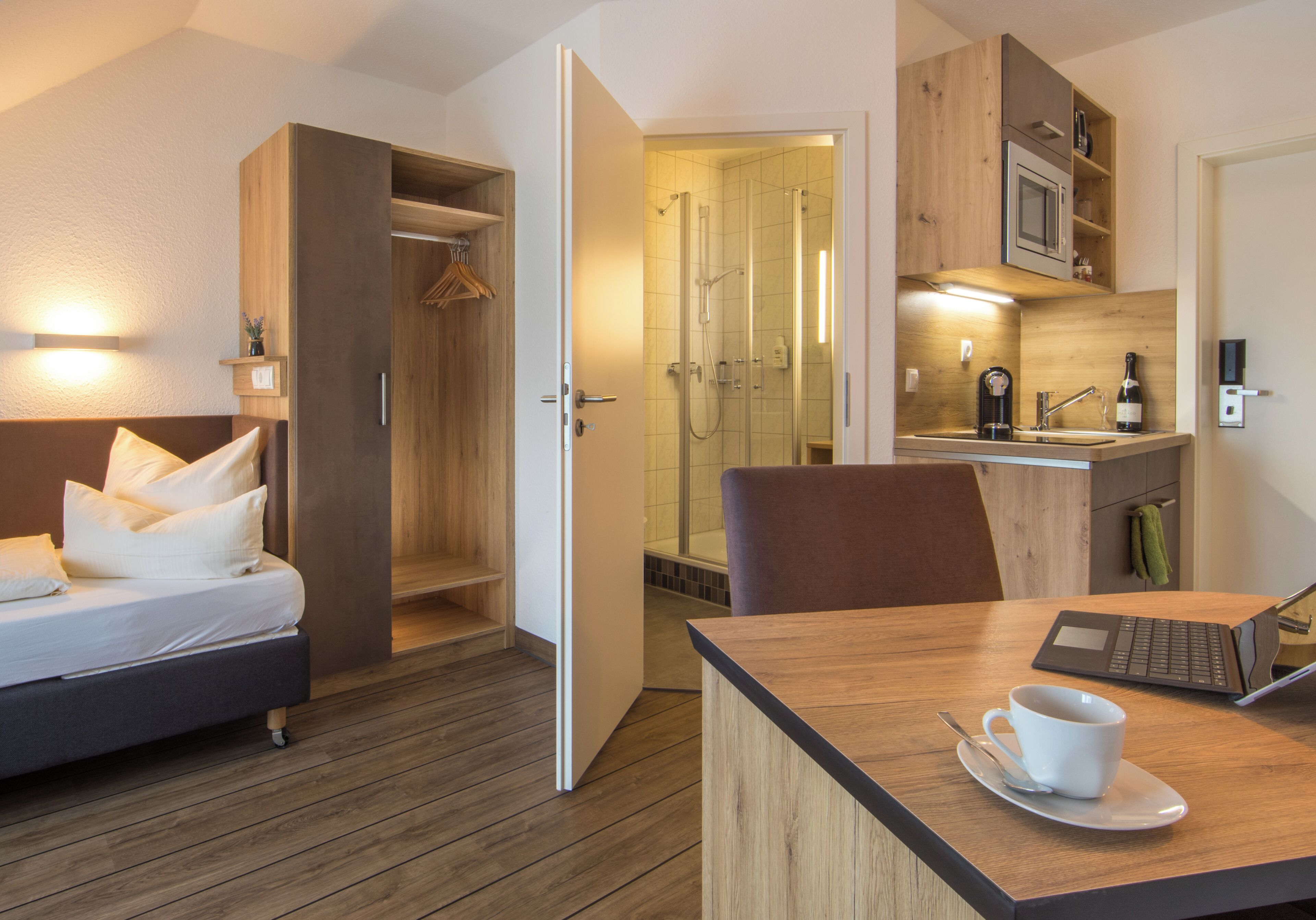 A hotel room with a bed, a desk with a laptop, a cup, and a shower area.