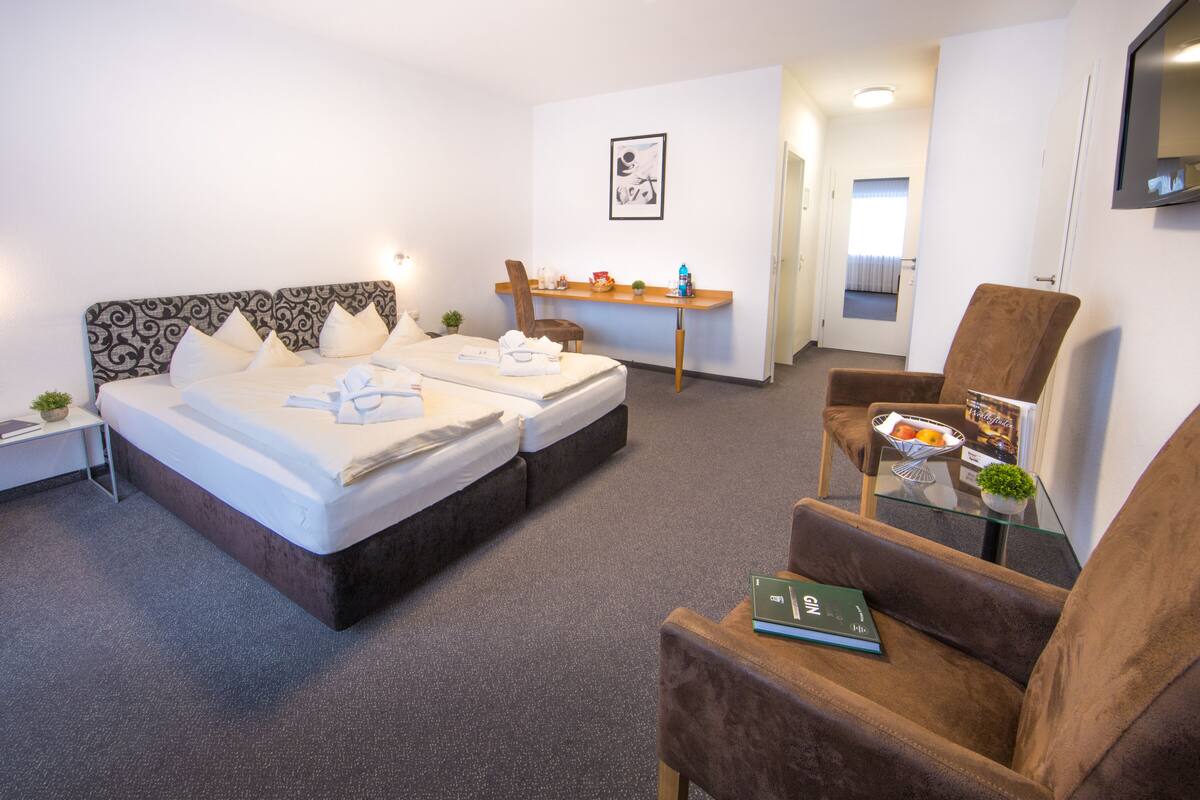A hotel room with a double bed, a small table with a plant, a chair, and a desk with a computer.