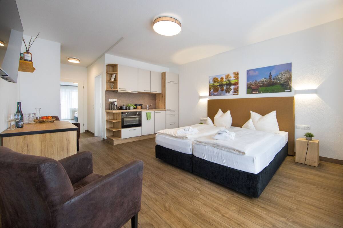 A modern hotel room with a large bed, a desk, and a kitchenette.