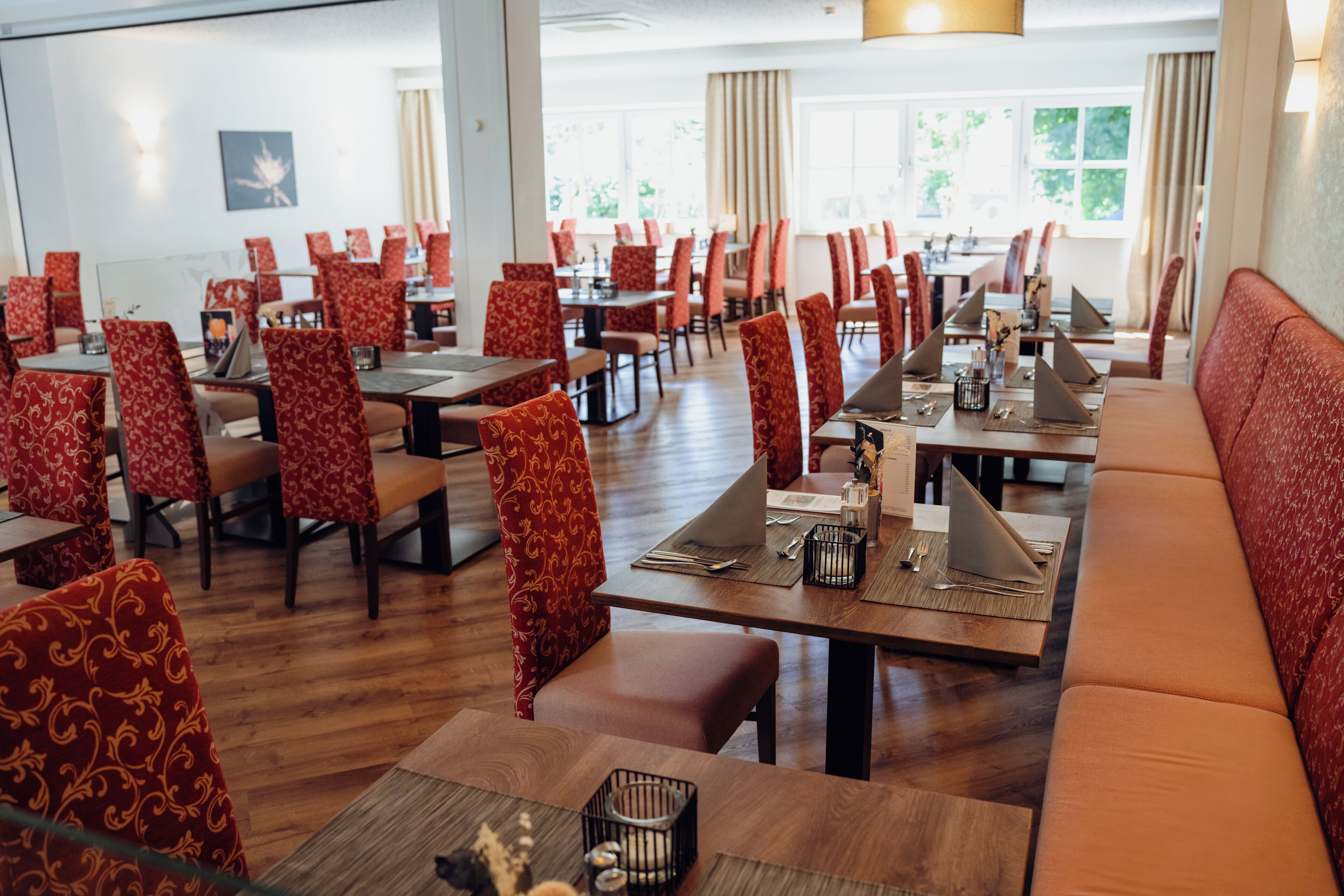 A restaurant with red patterned chairs and tables, wooden flooring, and a view of the outdoors through large windows.
