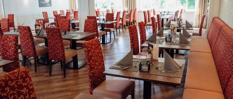 A restaurant with red patterned chairs and tables, wooden flooring, and a view of the outdoors through large windows.