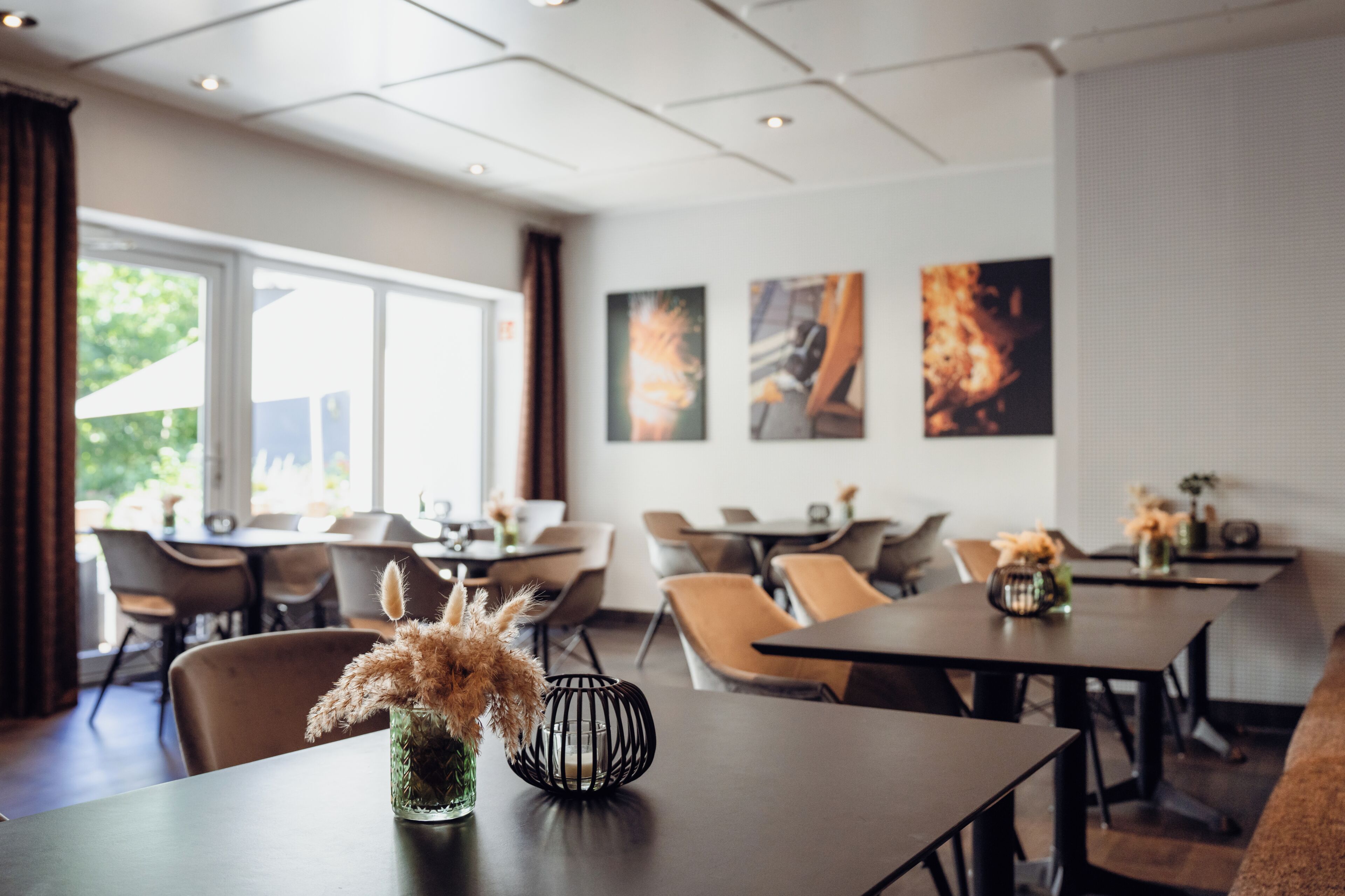 A modern dining area with tables and chairs, a vase with dried plants, and artwork on the wall.