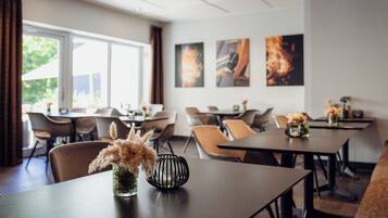 A modern dining area with tables and chairs, a vase with dried plants, and artwork on the wall.
