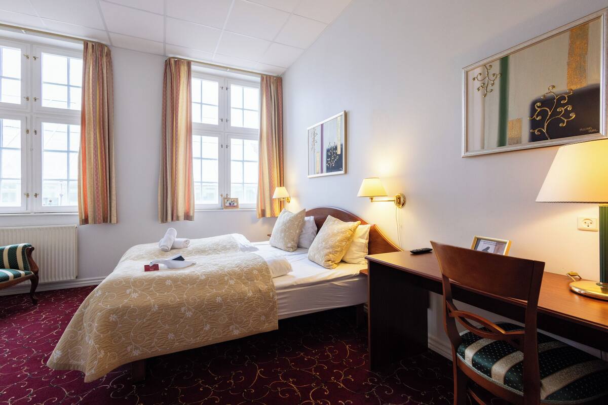 Ein Hotelzimmer mit einem Bett, einem Schreibtisch, einem Sessel, einem Fenster mit Vorhängen, einer Heizung und einer Wandlampe.
