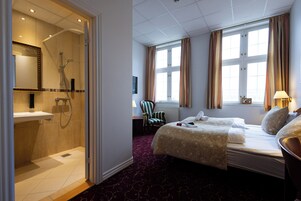 Quarto de hotel com chuveiro, cama com duas almofadas, uma cadeira e janela com cortinas.