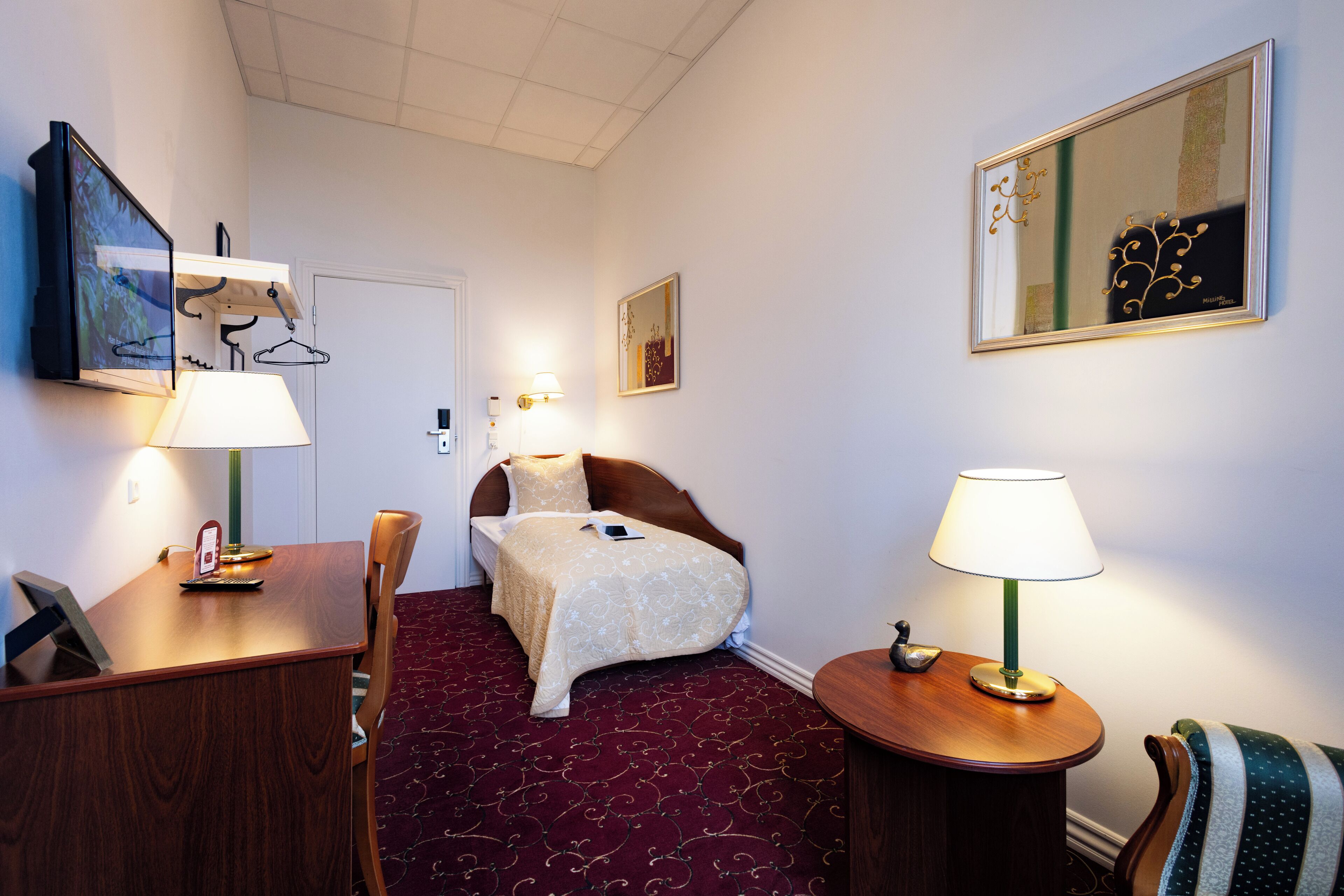 Quarto de hotel com cama, escrivaninha, cadeira, luminárias e obras de arte nas paredes.