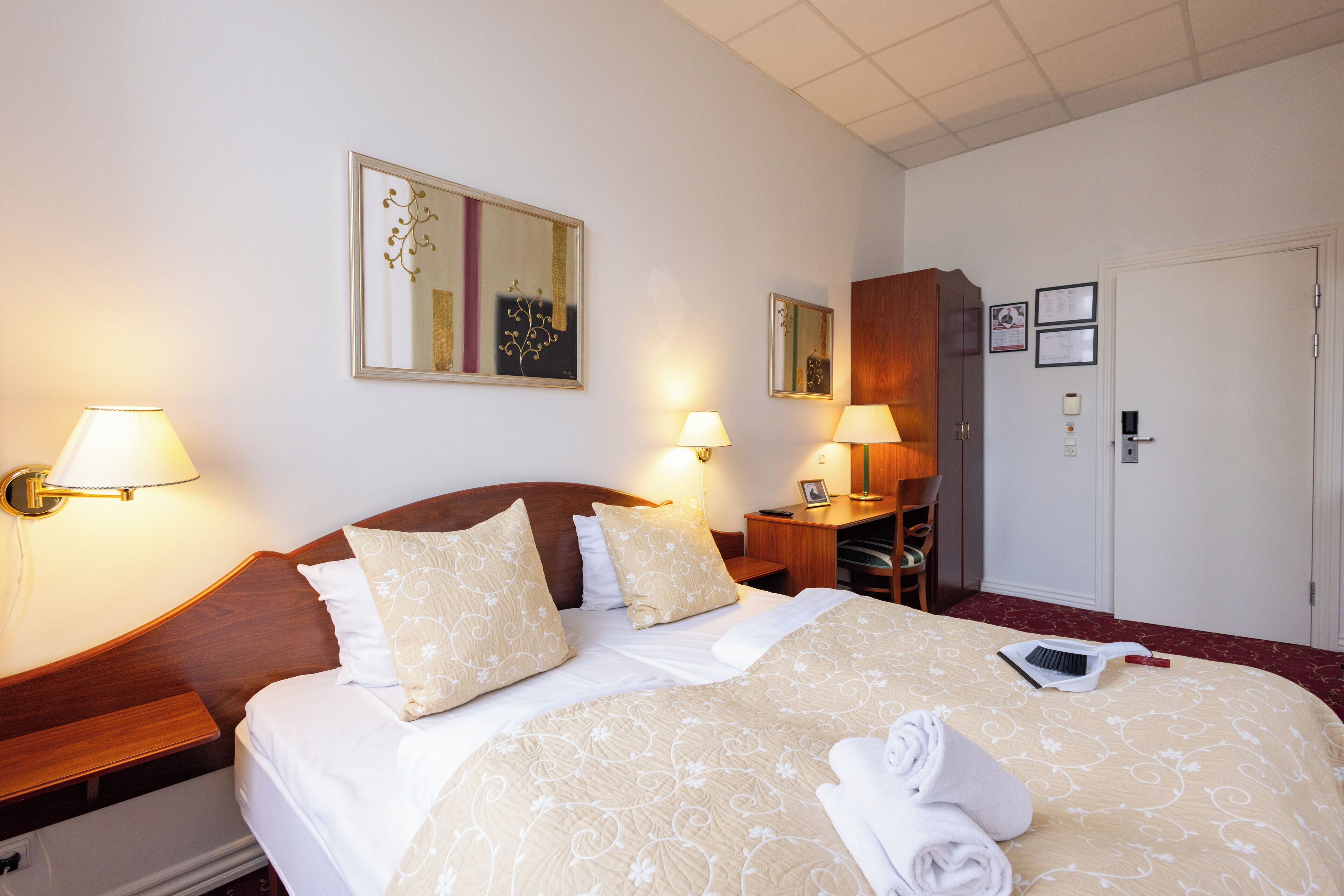 A hotel room with a neatly made bed, two bedside lamps, a wooden headboard, a desk with a chair, and a door with a handle.