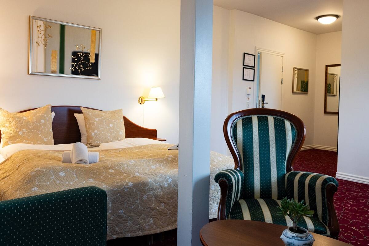 Ein Hotelzimmer mit einem ordentlich bezogenen Bett, einem Sessel, einem kleinen Tisch mit einer Vase und einem Spiegel an der Wand.