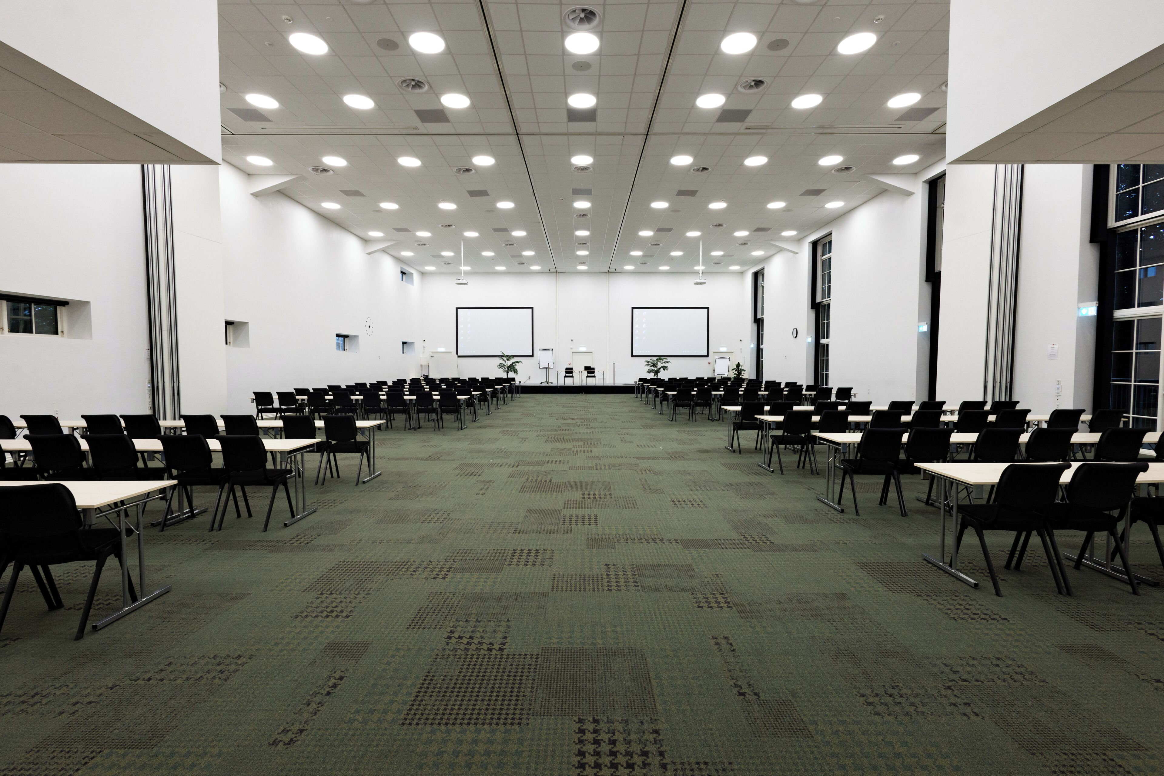 A conference room with rows of black chairs and tables, white screens, and a carpeted floor.