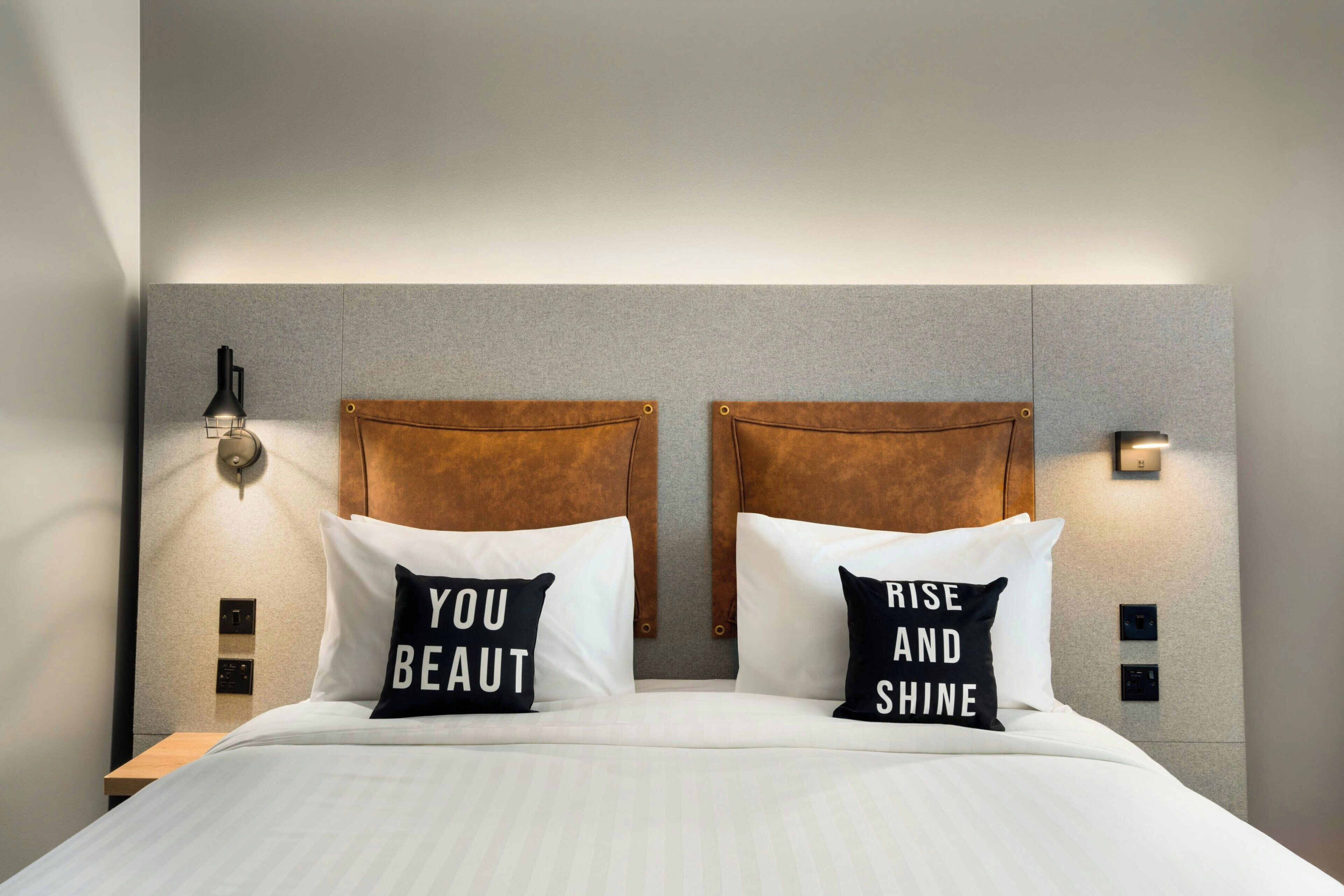 A hotel room with a bed, two pillows with motivational text, a headboard, and wall-mounted light fixtures.