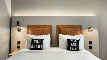 A hotel room with a bed, two pillows with motivational text, a headboard, and wall-mounted light fixtures.