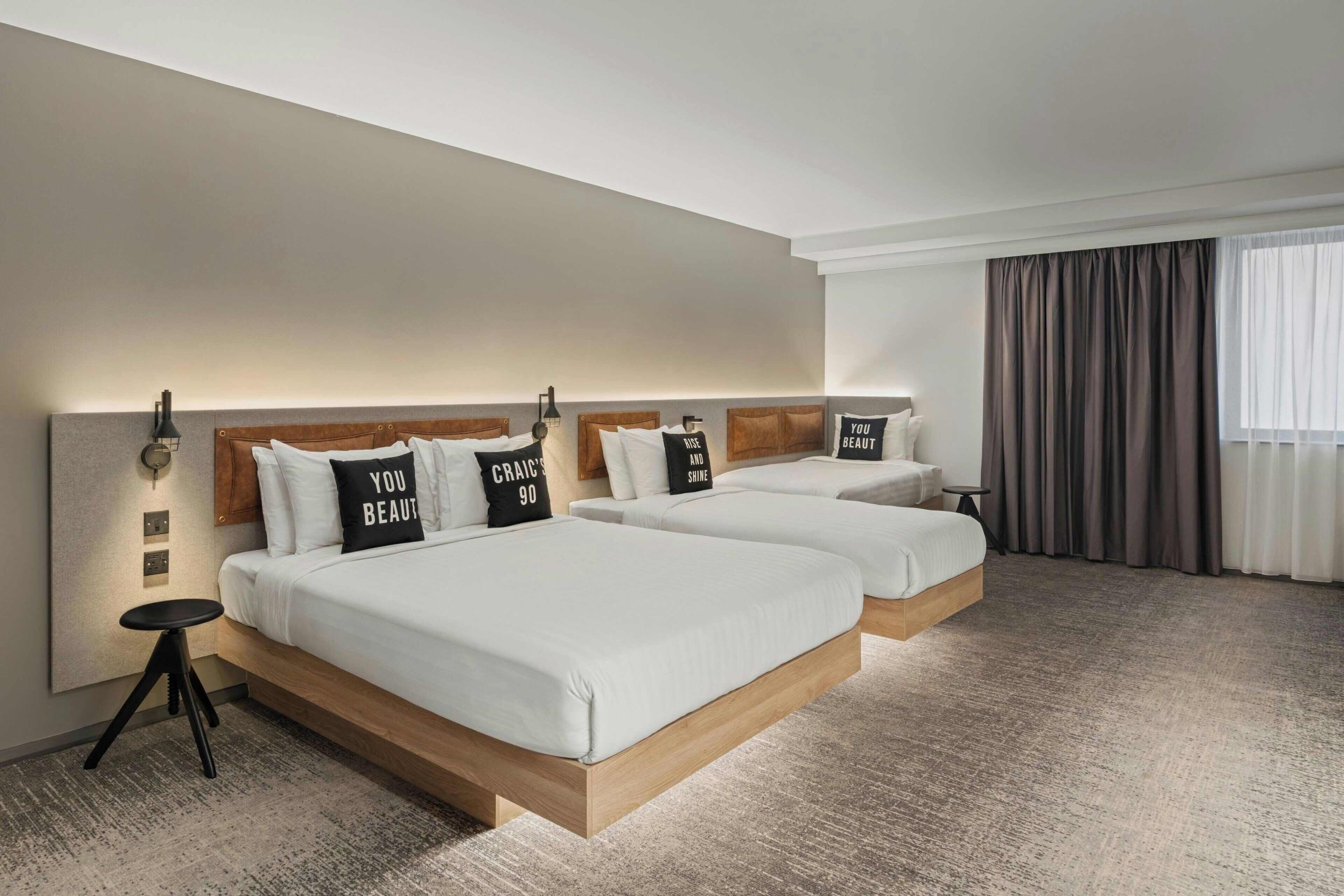 A hotel room with two beds, a small table, and a large window with curtains.