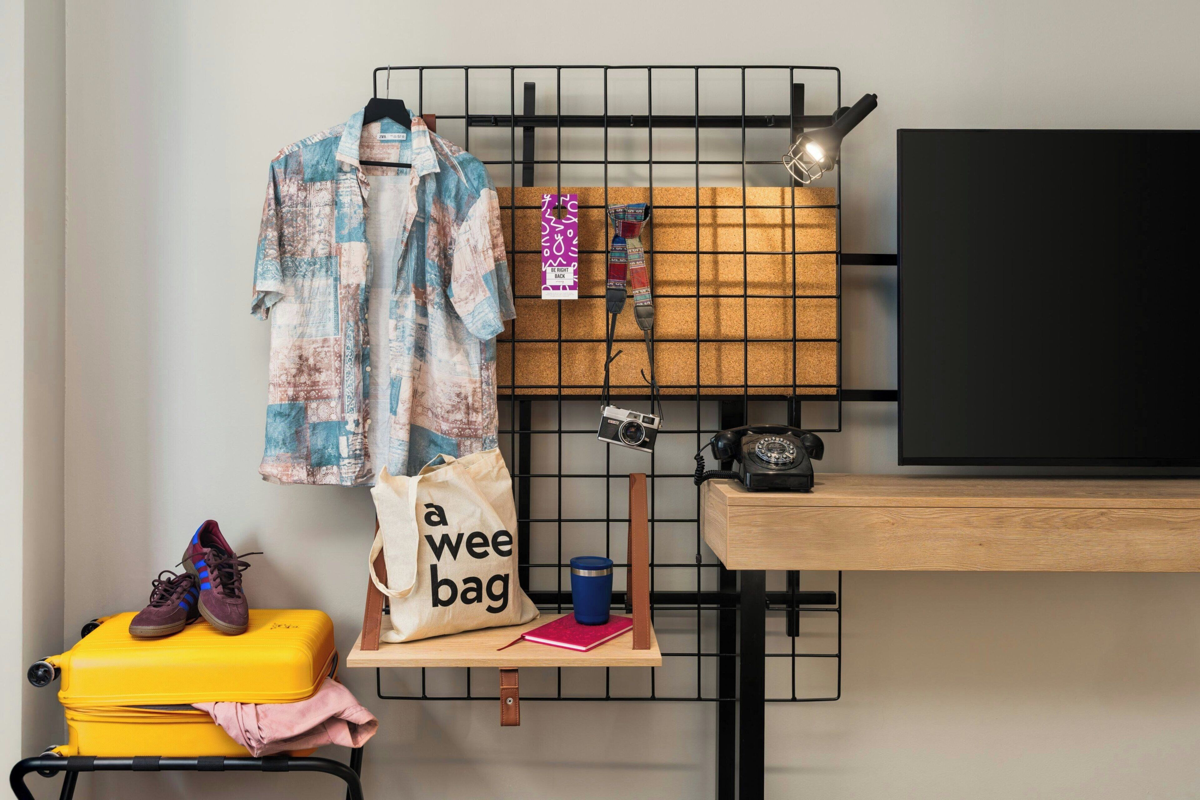 Una habitación con cama, escritorio, silla, un perchero con ropa y una cámara, además de una maleta.