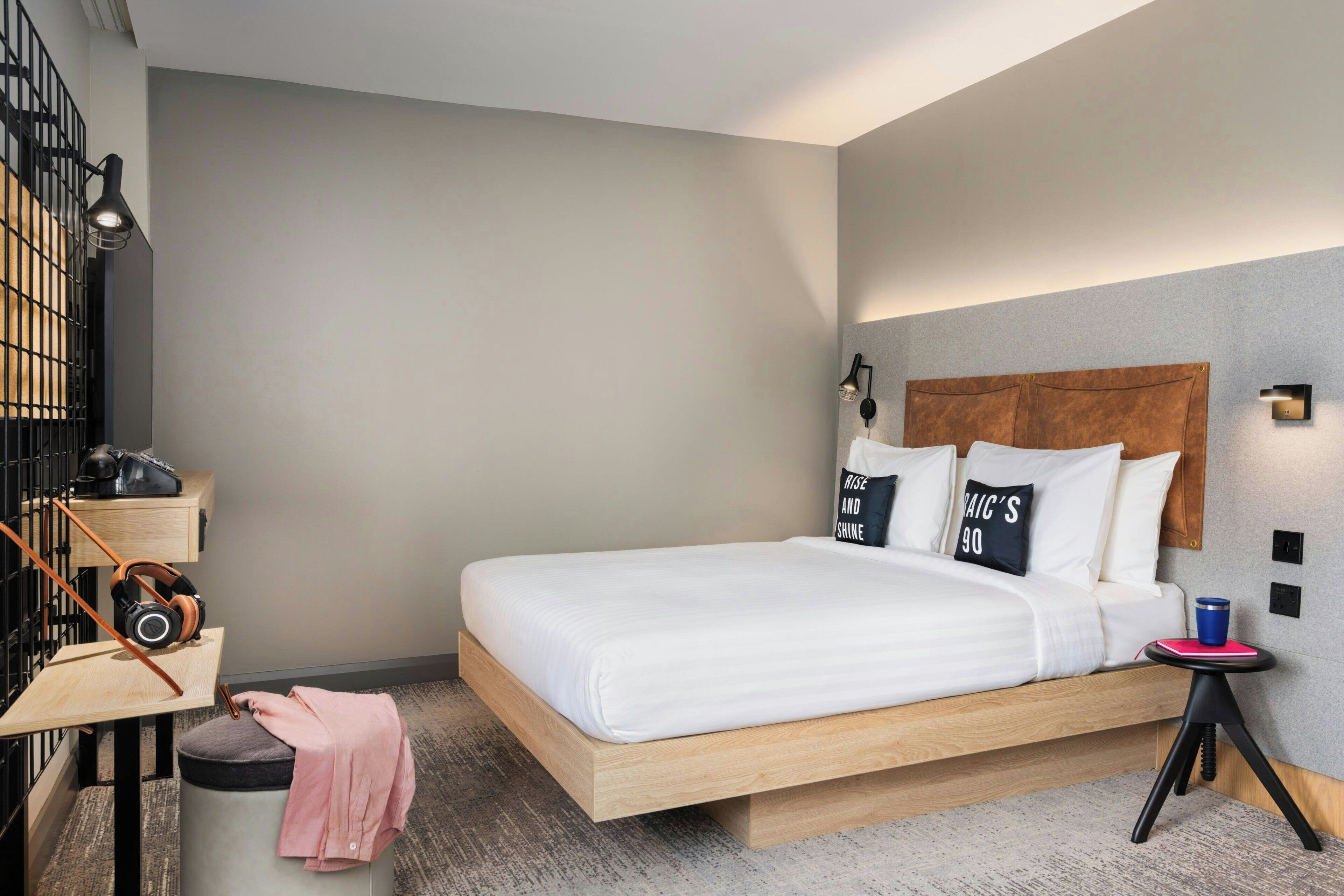 Une chambre d’hôtel moderne avec un lit en bois, une petite table avec un casque audio, une poubelle et une table de chevet avec une lampe.