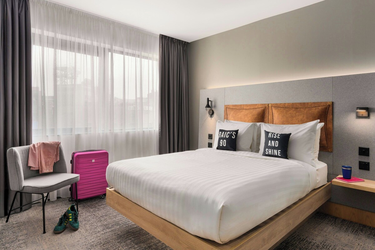 A modern hotel room with a large bed, a chair, a suitcase, and a nightstand.