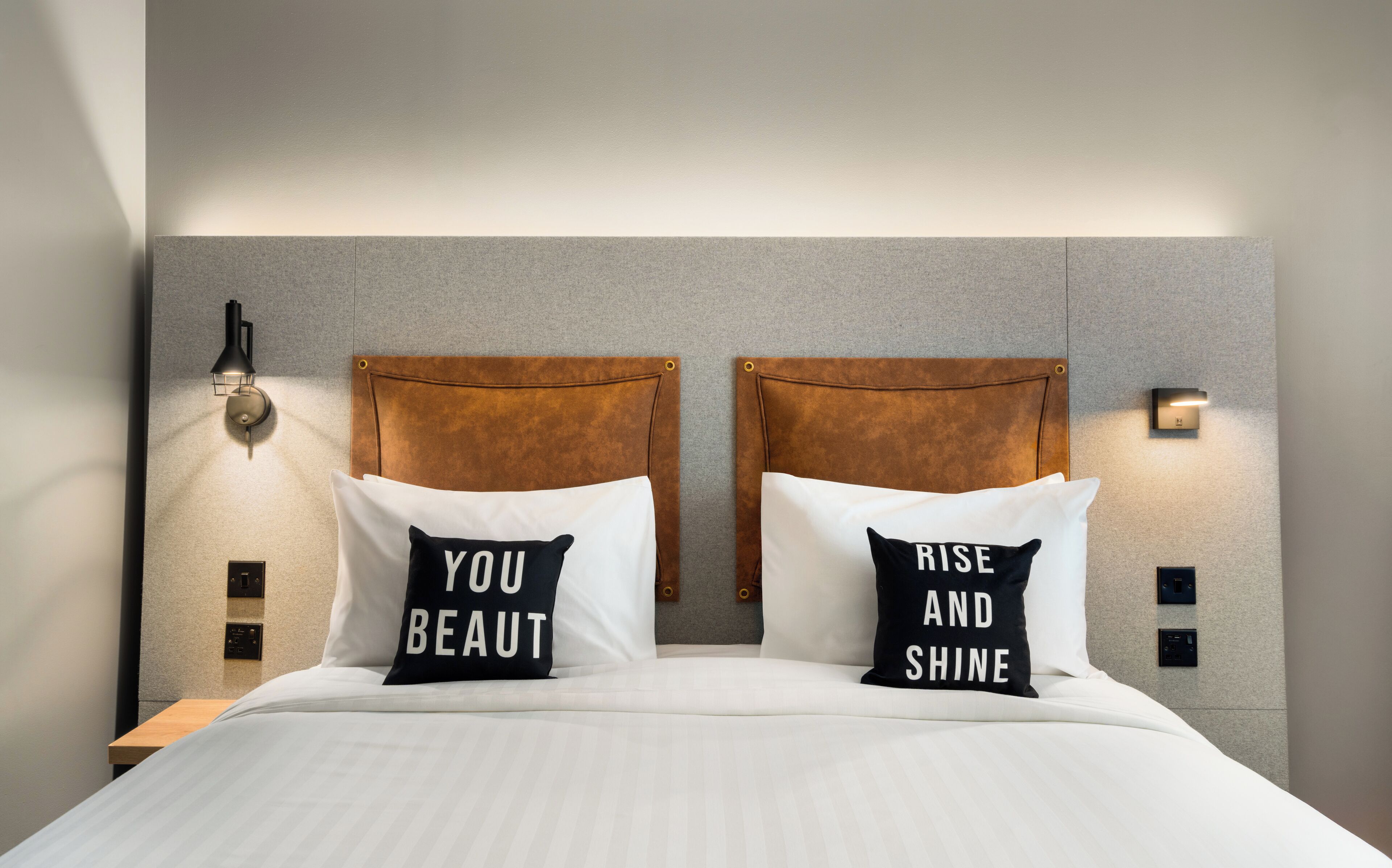 A neatly made bed with two pillows, one with "YOU BEAUTY" and the other with "RISE AND SHINE".